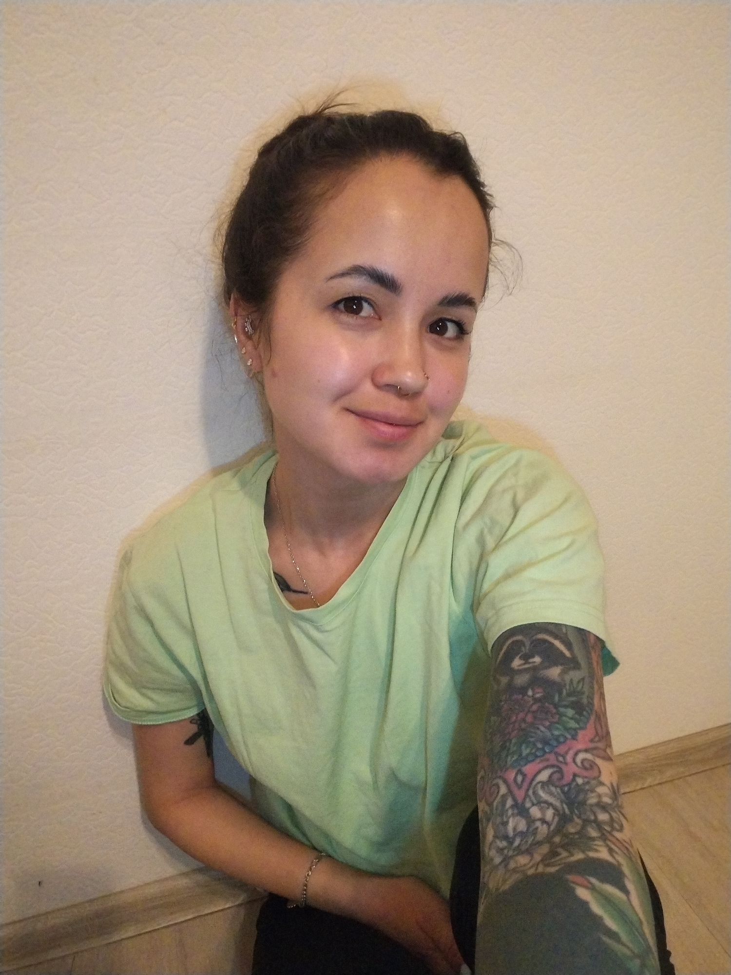 A young woman is depicted in a close-up portrait, seated on a light-colored wooden or laminate floor, leaning against a textured, off-white wall. She looks directly at the camera with a calm and gentle smile.
She has dark hair pulled back, with some loose strands framing her face. Her complexion is clear, and she has dark eyes. Notable facial features include a stud piercing in her left nostril. Her left ear is adorned with multiple piercings, including studs in the helix, tragus, and several lobe piercings.
She is wearing a simple, light green short-sleeved t-shirt and dark trousers or leggings. A thin silver chain necklace with a small pendant is visible around her neck, and a silver chain bracelet is on her left wrist.
Her arms are heavily tattooed. Her right arm features a full sleeve tattoo with intricate and colorful designs, including what appears to be a raccoon or similar animal at the top, alongside floral motifs and abstract patterns in shades of green, pink, blue, and black. Her left upper arm has a smaller, darker tattoo.
The setting is indoors, with a textured wall suggesting wallpaper or a painted textured surface, and a wooden floor. The lighting appears natural or well-distributed artificial light, indicating no specific time of day. No visible text is present in the image. The location context provided is Noviy, Russia.