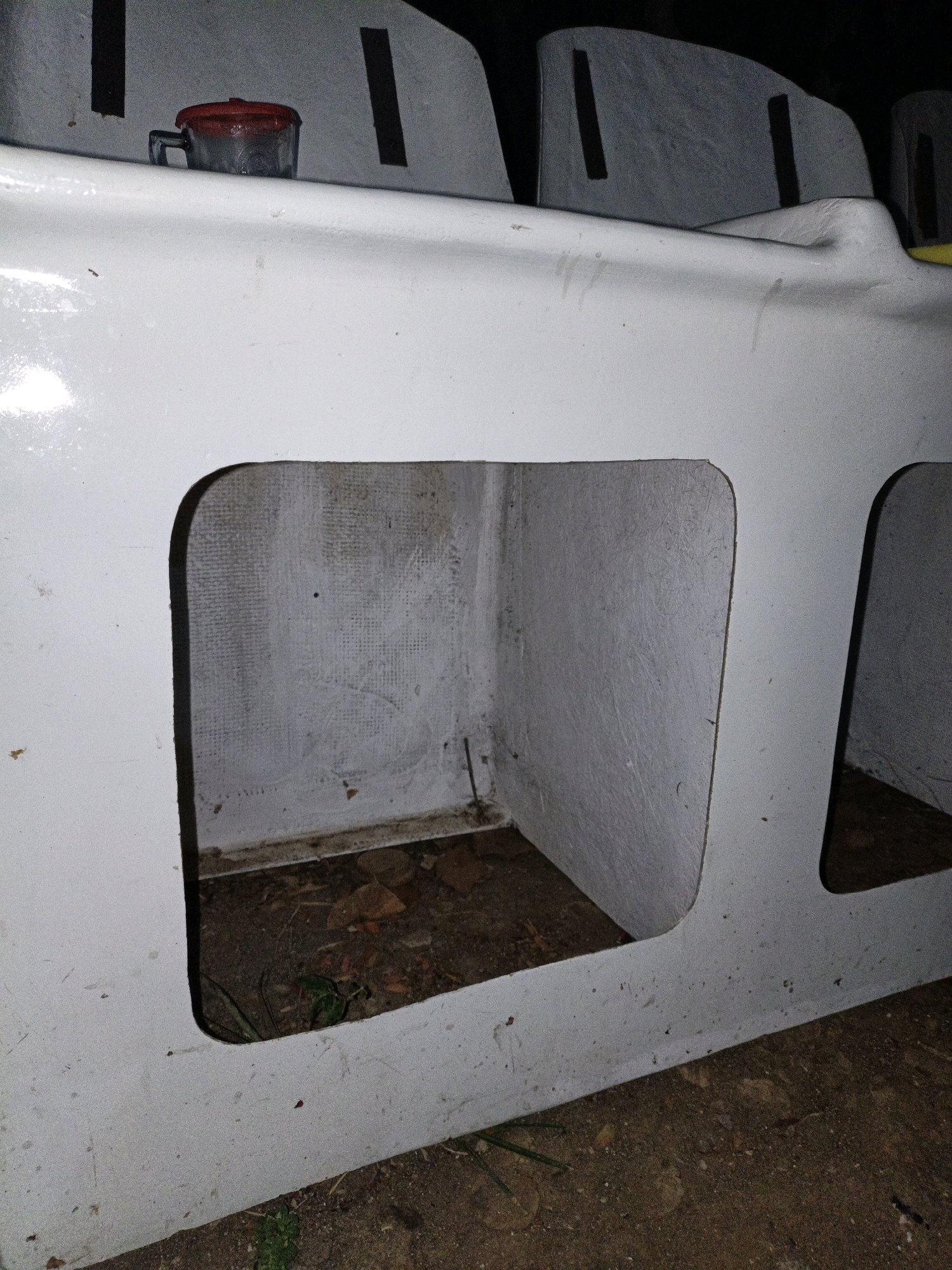 A close-up, low-angle exterior shot shows the side of a white, hollow structure with multiple cubby-like openings. The cubbies appear to be built into a larger, possibly modular, white unit. The texture of the interior surfaces of the cubbies is rough and gritty, with some evidence of dirt and debris. In the far left cubby, a small, clear glass container with a red lid is visible. The ground outside the structure is a mix of soil, small stones, and sparse green vegetation. The lighting suggests it is nighttime or a very dark interior space. The image does not provide any specific information about its location in the city of Cikal, Indonesia.