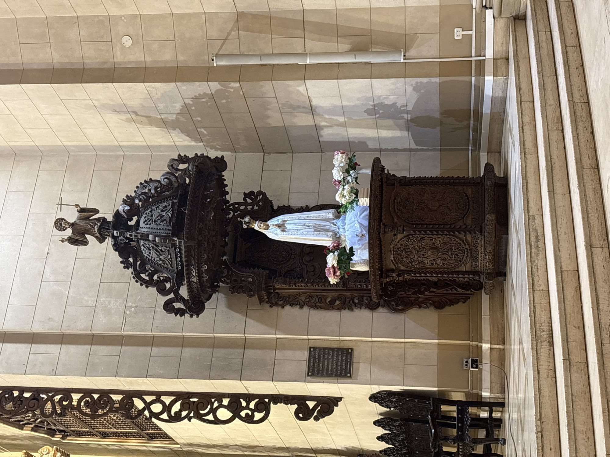 The image shows a religious statue of the Virgin Mary within a church, likely located in San Isidro, Peru. The statue is positioned on a carved wooden pedestal, adorned with flowers. Above the statue, on a darker, ornate wooden structure, is a smaller figure of Jesus Christ, holding a cross. The surrounding environment consists of stone walls and floor, with decorative elements like wrought iron grates and wooden chairs visible in the background. The lighting suggests an indoor setting, possibly with natural light filtering in from a window and artificial light sources. There is a plaque on the wall, as well as electrical outlets and speakers, indicating the presence of modern amenities within the historical setting. No individuals are visible in the scene.