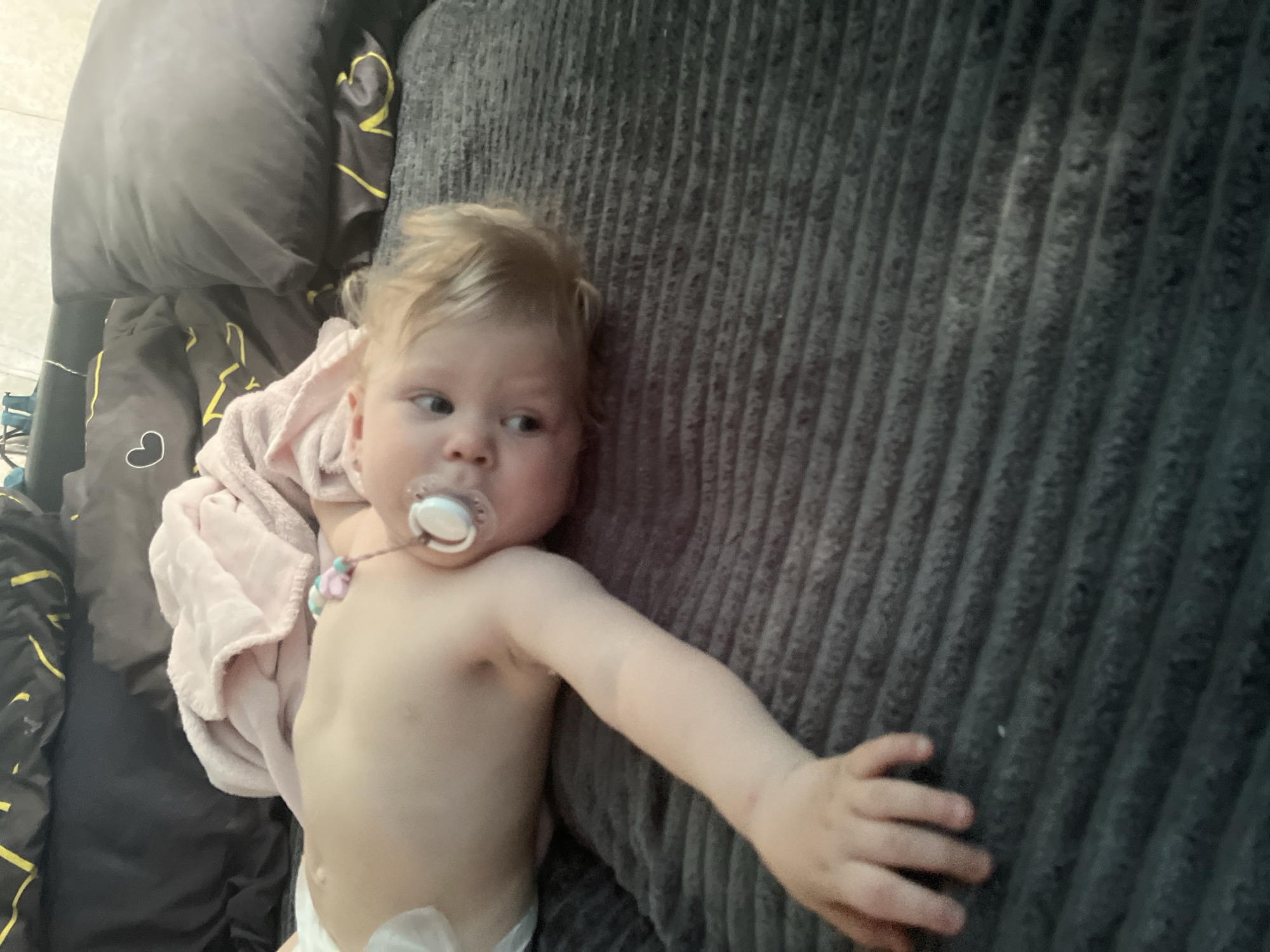 A baby with blonde hair and a pacifier is lying on a ribbed grey surface. The baby is wearing a diaper and a pink blanket is draped over its body. To the left of the baby are dark blankets with yellow stripes and a white heart outline. The baby's left arm is extended to the right, with its hand open. The scene appears to be indoors, with the lighting suggesting daytime. There is no visible text in the image. The overall mood is calm and relaxed.