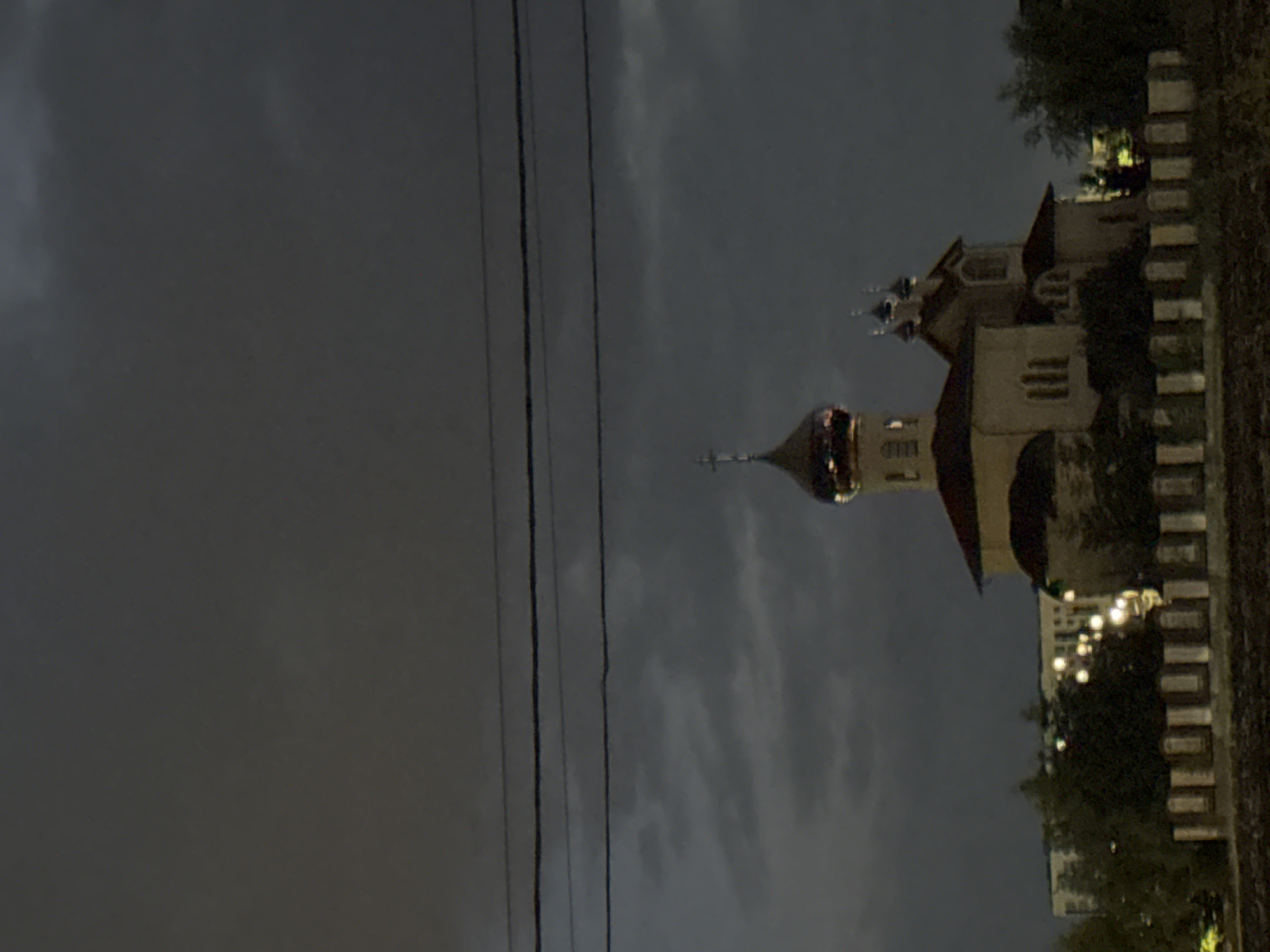 The image depicts an Orthodox church with multiple domes, a prominent bell tower, and several smaller towers, situated in Mikhaylovsk, Russia. The church is constructed of light-colored material, with a red-tiled roof. It is dusk or night, with a dark, cloudy sky dominating the upper portion of the image. Several dark power lines are visible in the sky above the church. In the background, buildings and streetlights are illuminated, suggesting the presence of a residential or urban area. In the foreground, a low wall or fence runs horizontally across the image, with visible vertical elements that appear to be grave markers or decorative elements. Trees are present to the left and right of the church, with some appearing to be illuminated by distant lights. The overall mood of the image is somber and atmospheric, due to the low light conditions and the overcast sky.