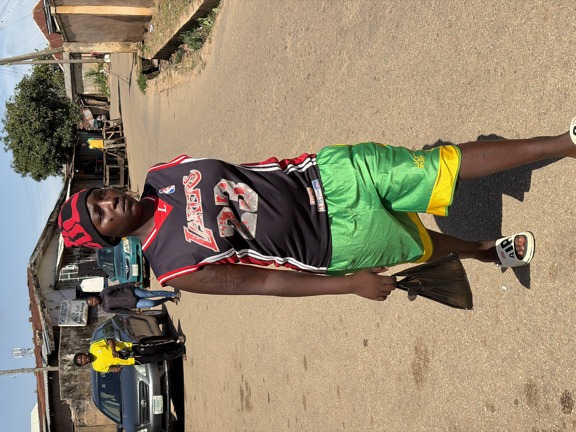 A medium shot captures a person walking on a dirt road in Jos, Nigeria. The individual is wearing a red and black beanie, a black basketball jersey with red and white trim, and bright green shorts with yellow trim. They are carrying a black plastic bag in their right hand and walking towards the viewer's right. The person has some markings on their face.
In the background, two cars are parked on the left side of the road. The car closest to the viewer is silver, and the other is a light green sedan. A man in a yellow shirt and black pants is standing next to the silver car, looking towards the viewer. Behind the cars, there are buildings with corrugated iron roofs and some market stalls. The road surface is uneven and dusty. The sky is clear and blue. The overall scene depicts a common street view in a Nigerian city.