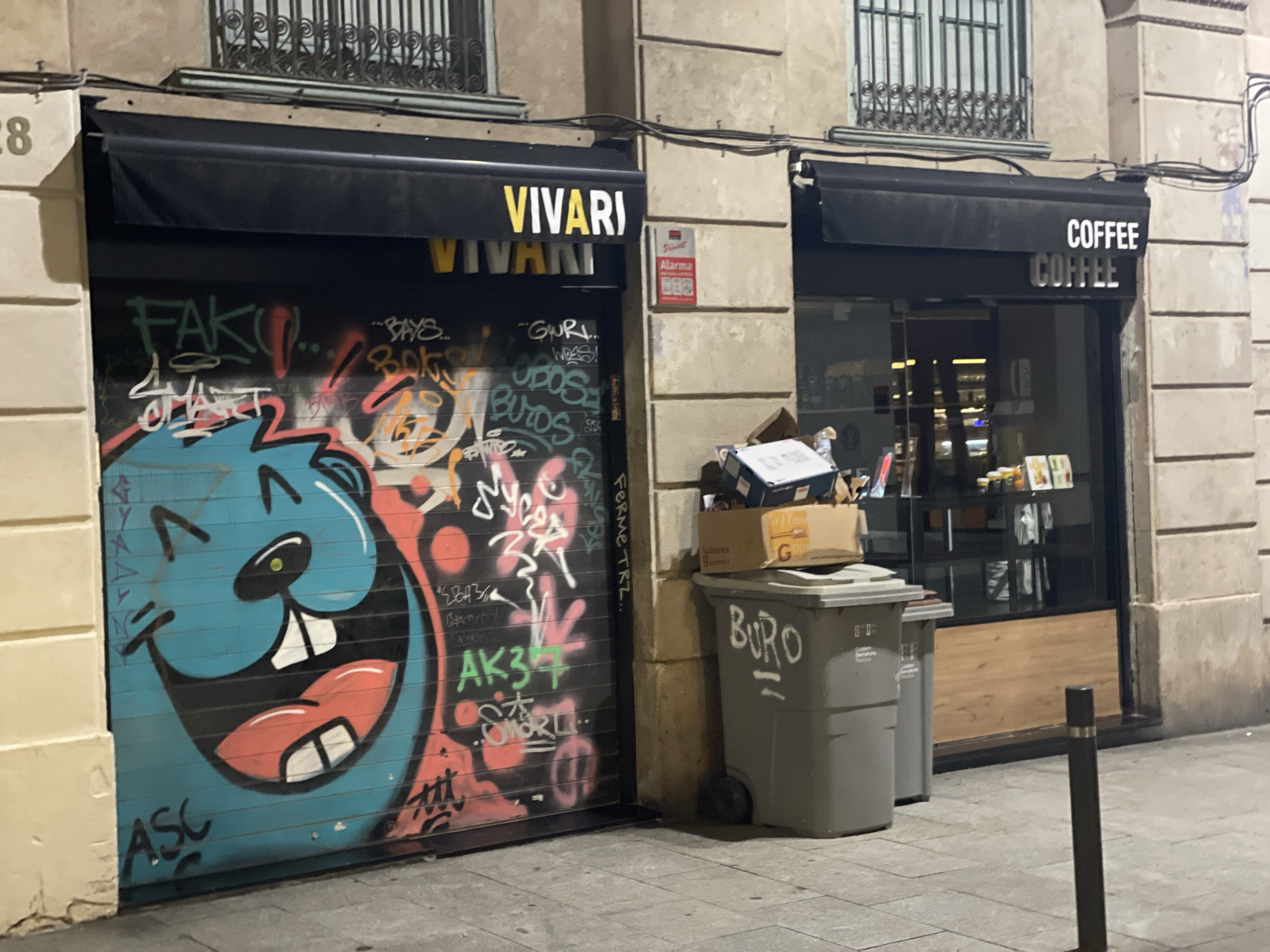 The image shows a street scene in Barcelona, Spain, at night. The focus is on the storefronts of two businesses. The left storefront is a roller shutter, heavily graffitied with a large, cartoonish blue character's face and various taggings. Above the shutter, the word "VIVARI" is displayed in yellow and white lettering on a black awning. To the right of this, a red alarm sign is visible on a stone building facade.
The right storefront is a cafe with a glass window displaying merchandise and a wooden panel below. A black awning above the window has "COFFEE" written in white lettering. Next to the coffee shop entrance, there are two large gray trash bins, one stacked with cardboard boxes. The ground is paved with gray stones. The building facades are made of light-colored stone. The overall impression is an urban street at night, with a contrast between the vibrant graffiti and the commercial establishments.