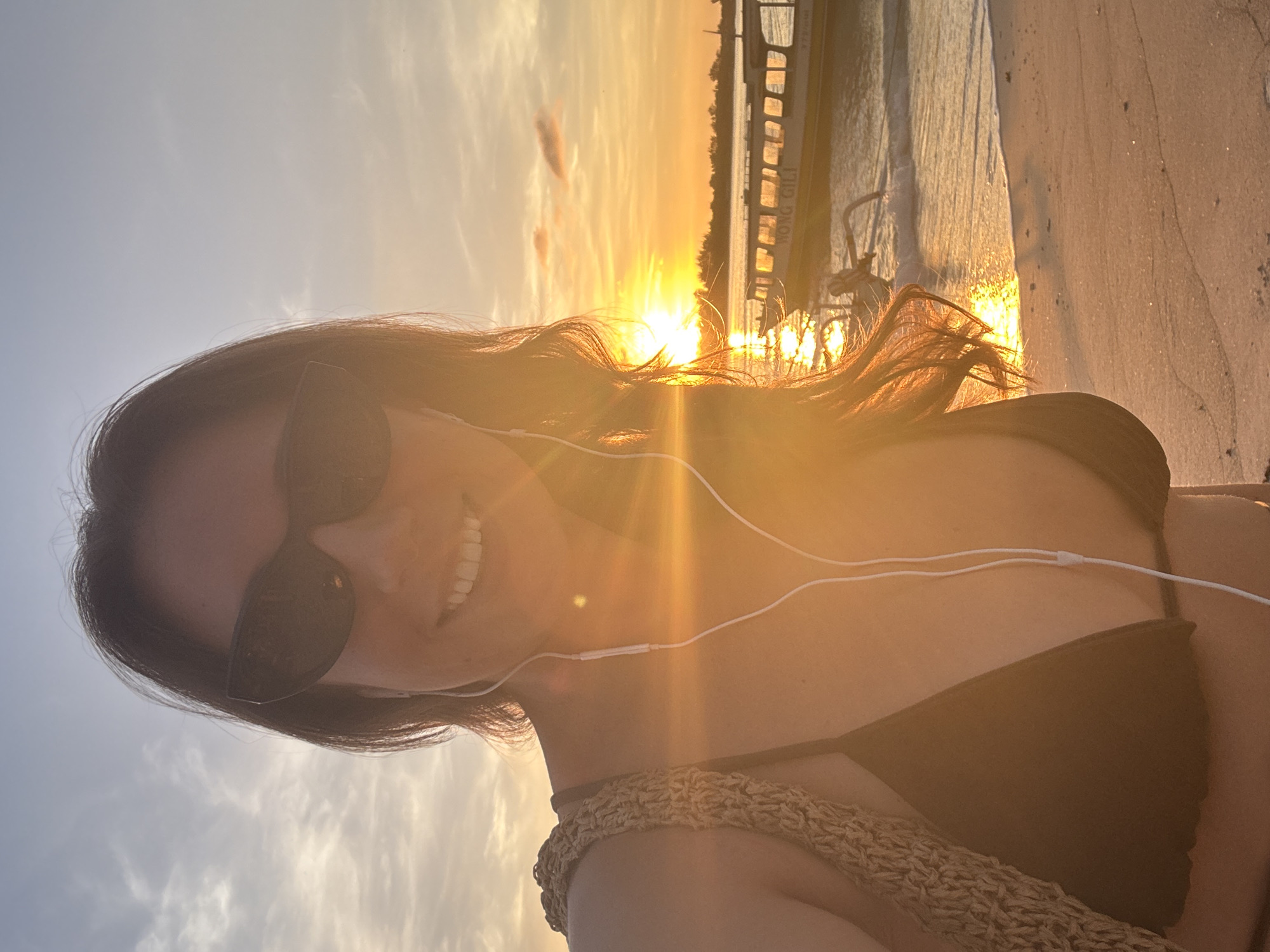A woman with dark hair and sunglasses is taking a selfie on a beach at sunset. She is wearing a black bikini top and has white earphones in her ears. The sun is setting over the water, and there appears to be a pier or dock in the background. The location is likely in Unknown, Indonesia. The beach sand is visible in the foreground.