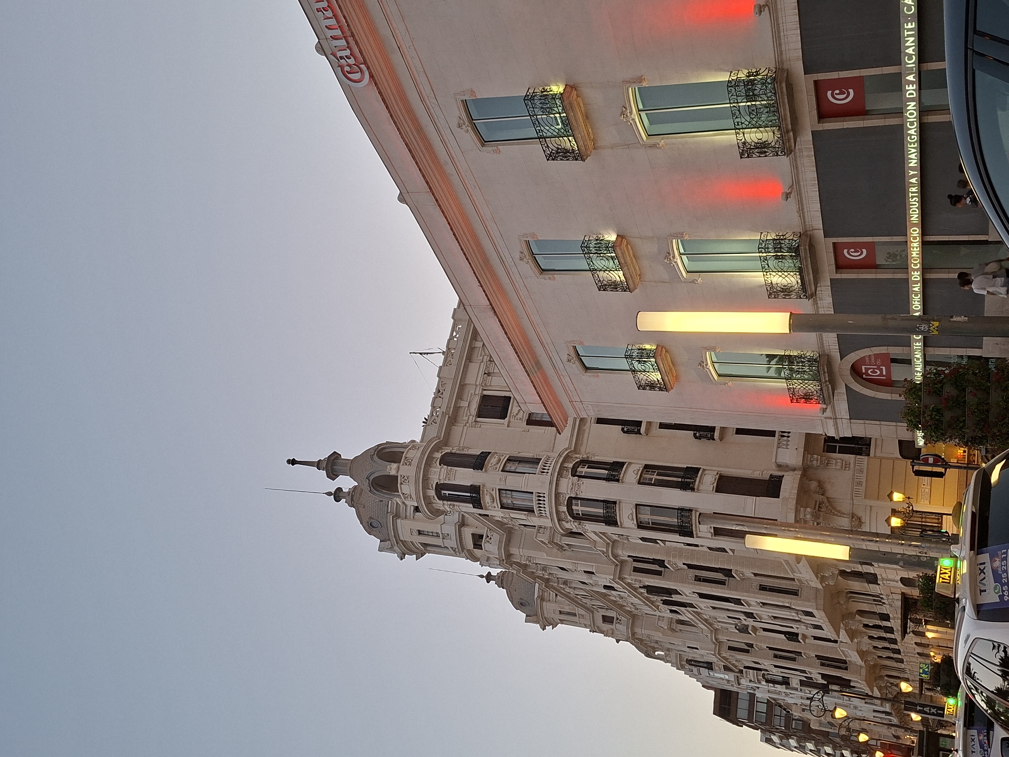 The image captures a street scene in Alicante, Spain, likely near the city center, given the architecture and the presence of a "Caja de Ahorros del Mediterráneo" (CAM) bank, suggested by the prominent "CAM" signage. The perspective is from street level, looking up at a row of ornate buildings and a modern bank building. The sky is a soft, pale blue, indicating dusk or early morning light.
The building on the right is a light-colored, multi-story structure with large, rectangular windows, each featuring decorative black iron balconies. Red lights are projected onto the facade below the windows, creating a striking visual effect. The CAM logo, a red circle with a white "C" inside, is visible on two levels of the building. A long, illuminated sign below the windows reads "CÁMARA OFICIAL DE COMERCIO INDUSTRIA Y NAVEGACIÓN DE ALICANTE".
To the left of the CAM building is a much older, elaborate white building with multiple turrets, domes, and balconies, characteristic of Beaux-Arts or similar late 19th/early 20th-century European architecture. This building has many windows and intricate decorative details.
In the foreground, several white taxis with illuminated green "TAXI" signs are visible, indicating a busy street. A tall, cylindrical light fixture with a bright yellow glow stands in front of the CAM building. Two individuals, one wearing a white shirt and the other a dark shirt, are walking on the sidewalk in front of the CAM building.
The overall impression is a blend of historic and modern urban architecture illuminated by artificial lighting as daylight fades.