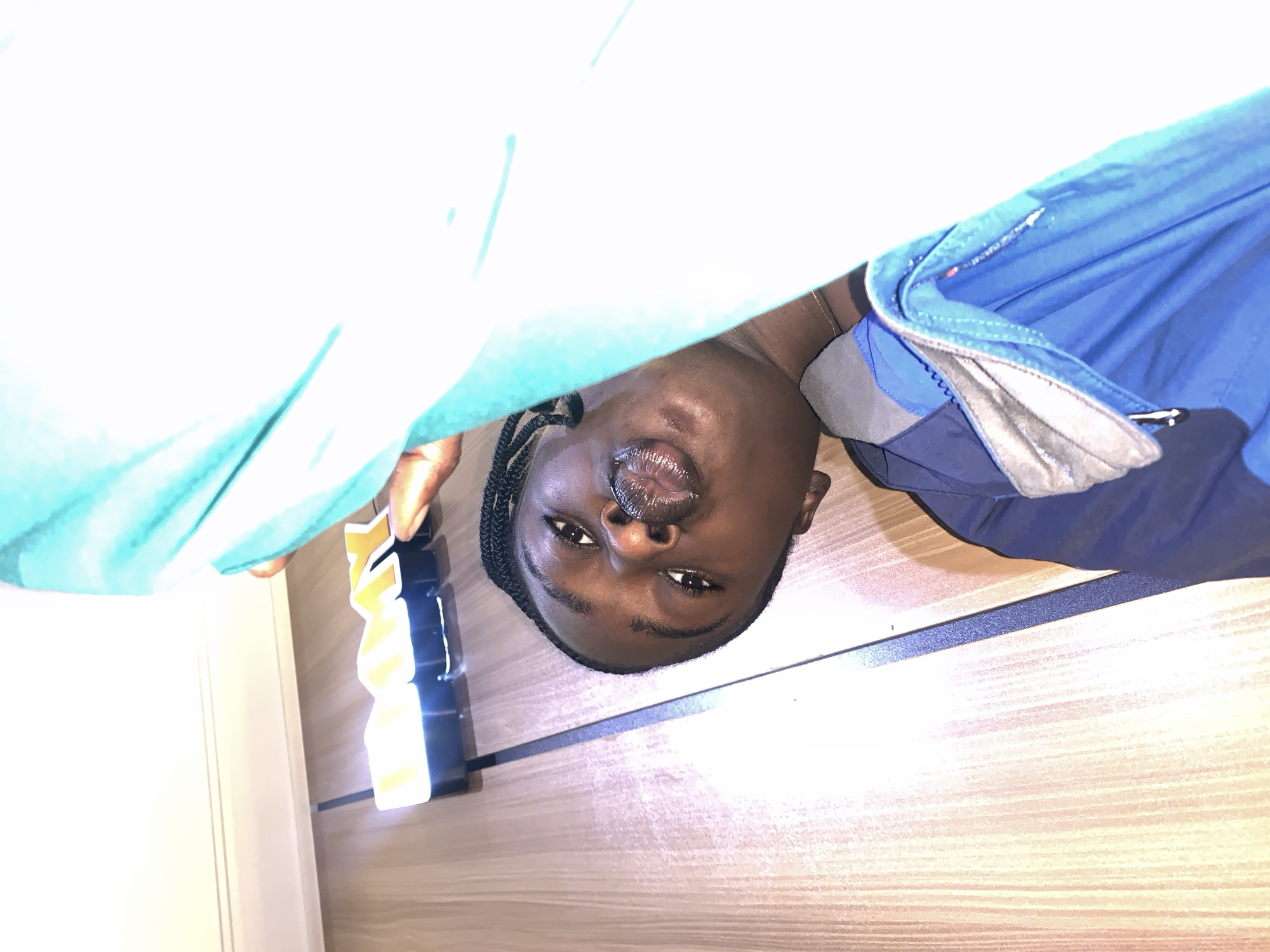 A person is lying on a wooden surface, with their head near a lit sign that spells "DMY". The person is looking into the camera with their lips puckered. They are wearing a blue jacket. The background is mostly white, with some of the person's body and clothing visible. The lighting is bright and casts a glow on the sign. The scene appears to be indoors.