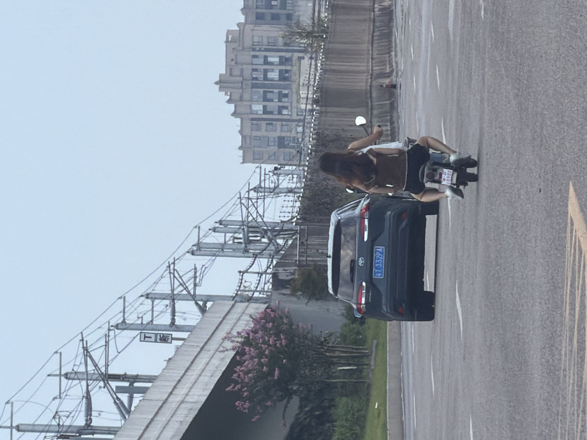 Two people ride a scooter on a road in Jiangjin, China. The person in front is wearing a white tank top and denim shorts, and the person behind is wearing a brown vest over a white top and denim shorts. The scooter has a white license plate with red characters. The road is paved with white lane markings. To the left of the road is a dark-colored SUV with a blue license plate reading "QT 332PA." A large bridge structure with overhead power lines is visible to the left, and a multi-story building with numerous windows is in the background on the right. The sky is overcast.