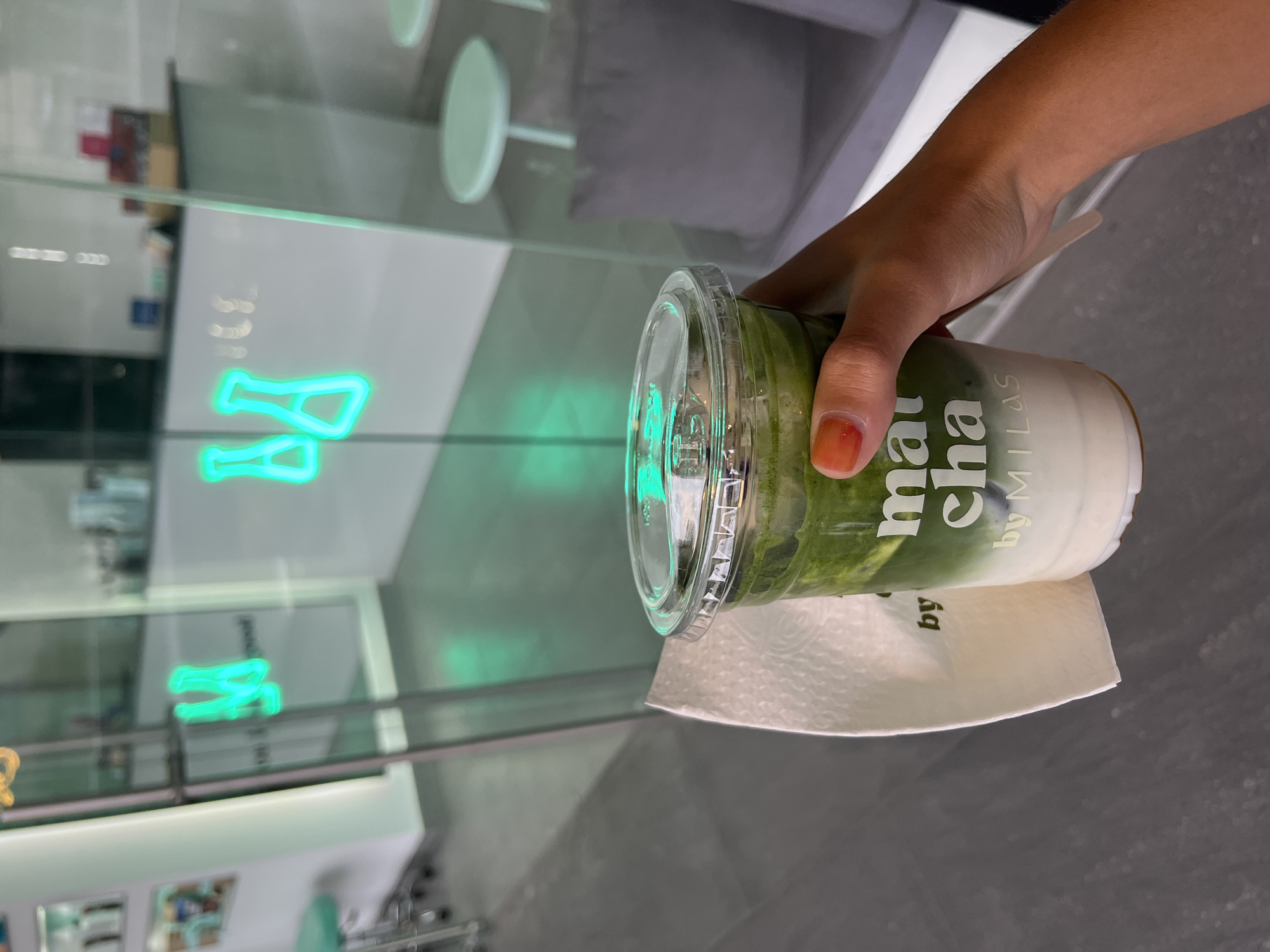 A person is holding a clear plastic cup containing a green liquid and a white layer at the bottom. The cup is sitting on a white paper napkin. The hand holding the cup has a painted nail. The background shows a blurred interior with a mirrored surface, possibly a window or partition. A neon sign is reflected in the surface. The location is unclear from the image. The city is mentioned in the prompt but not depicted in the image.