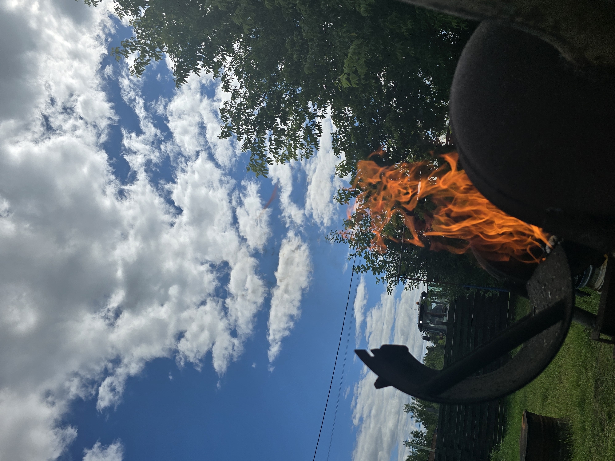 In Ермолаево, Russia, under a bright blue sky dotted with white, fluffy clouds, a fire burns vigorously in a metal structure, likely a grill or brazier. The flames, a vibrant orange and yellow, leap upwards. In the foreground, a dark, curved metal object obscures a portion of the fire and the surrounding scene. To the left of the fire, a dark silhouette of a hand with an extended index finger is visible against the sky. In the background, a dark wooden fence runs horizontally, and behind it, a green tractor is parked on grassy terrain. Trees with lush green foliage flank the scene on the right and in the distance. A dark, cylindrical object, possibly a barrel, rests on the grass to the left. Power lines are strung across the sky.