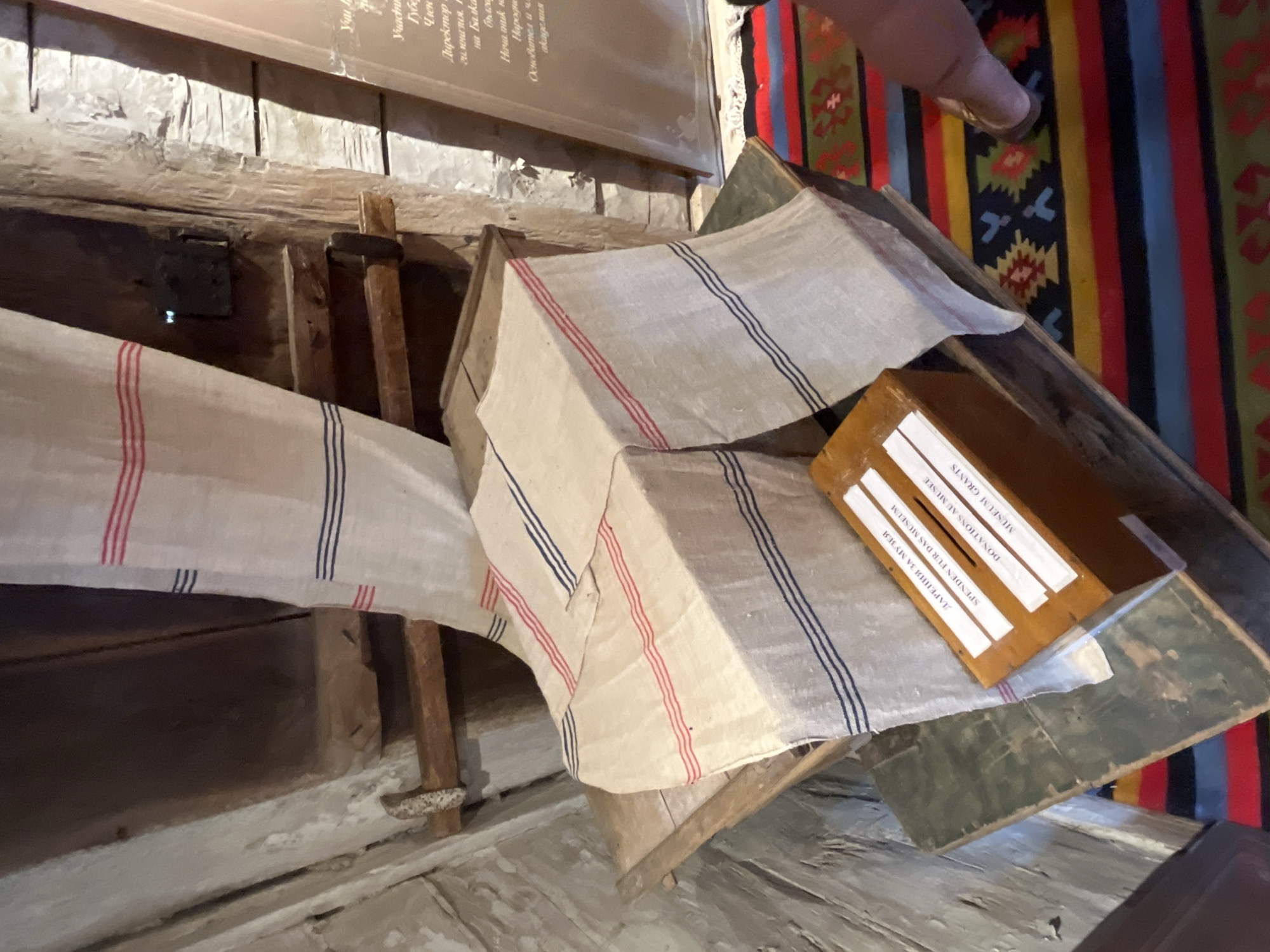 The image depicts an interior scene, possibly within the city of Zheravna, Bulgaria, given the context of the provided crops. The primary focus is on a wooden donation box, intricately designed with multiple labels in different languages. One label reads "ДАРЕНИЯ ЗА МУЗЕЯ" in Cyrillic script, suggesting "Donations for the Museum." Below this, "SPENDEN FUR DAS MUSEUM" is visible in German, and further down, "DONATIONS AU MUSEE" in French, and "MUSEUM GRANTS" in English. The donation box is positioned on a wooden surface covered by a piece of striped fabric, featuring red and blue stripes on a natural linen background. This fabric is draped over a rustic wooden table or display. In the background, rough wooden beams and walls suggest an old or traditional structure. A colorful, patterned rug with geometric designs lies on the floor beneath the display. A person's foot, wearing a sandal, is visible stepping onto the rug on the right side of the frame. The overall impression is of a historical setting where visitors are encouraged to contribute to the preservation of cultural heritage.