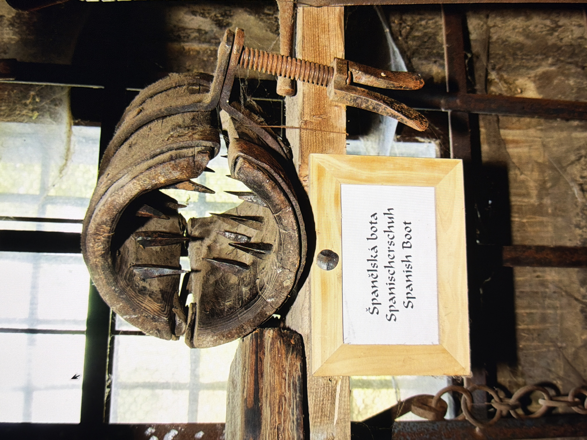 The image shows a display of a medieval torture device, known as the Spanish boot, in what appears to be a museum or historical exhibit. The device is made of wood and has several spikes on the inside, designed to crush a victim's leg. It is mounted on a wooden shelf, and a sign in front of it provides its name in Czech, German, and English. The background suggests a rustic setting, possibly an old building with wooden beams and a window. The lighting is dim, creating a somber atmosphere. There are no people visible in the image, and no actions are taking place. The context suggests this is a historical artifact being presented for educational purposes. This object is not located in the city of Kyiv, Ukraine.
