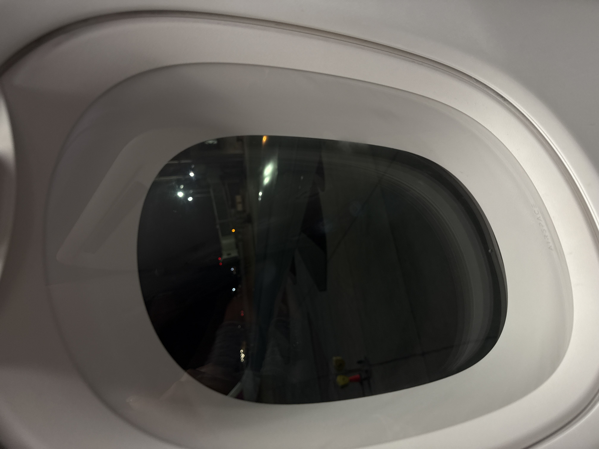 The image provides an interior view of an aircraft window. The window is oval-shaped and set within clean, light-grey or white cabin paneling, which features smooth, curved lines. The glass of the window is highly reflective, dominating the scene and obscuring a clear view of the external environment.
Within these reflections, several bright, artificial lights are visible, scattered across the dark background. These lights, along with the indistinct, shadowy shapes reflected, suggest the aircraft is either inside a brightly lit hangar or parked near an illuminated building during nighttime. Partial reflections of what appears to be a person, possibly the photographer's arm or leg, are visible on the left side of the window. In the lower right of the reflection, small yellow and red objects can be discerned, resembling equipment or cables.
The aircraft's interior paneling appears modern and well-maintained. Faint text is visible on the right side of the window frame, reading "AIRSPACE," indicative of the Airbus cabin design. The overall scene depicts a stationary aircraft, potentially on the ground at an airport in Yerevan, Armenia. There is no specific activity or event taking place, conveying a tranquil moment from inside the cabin.