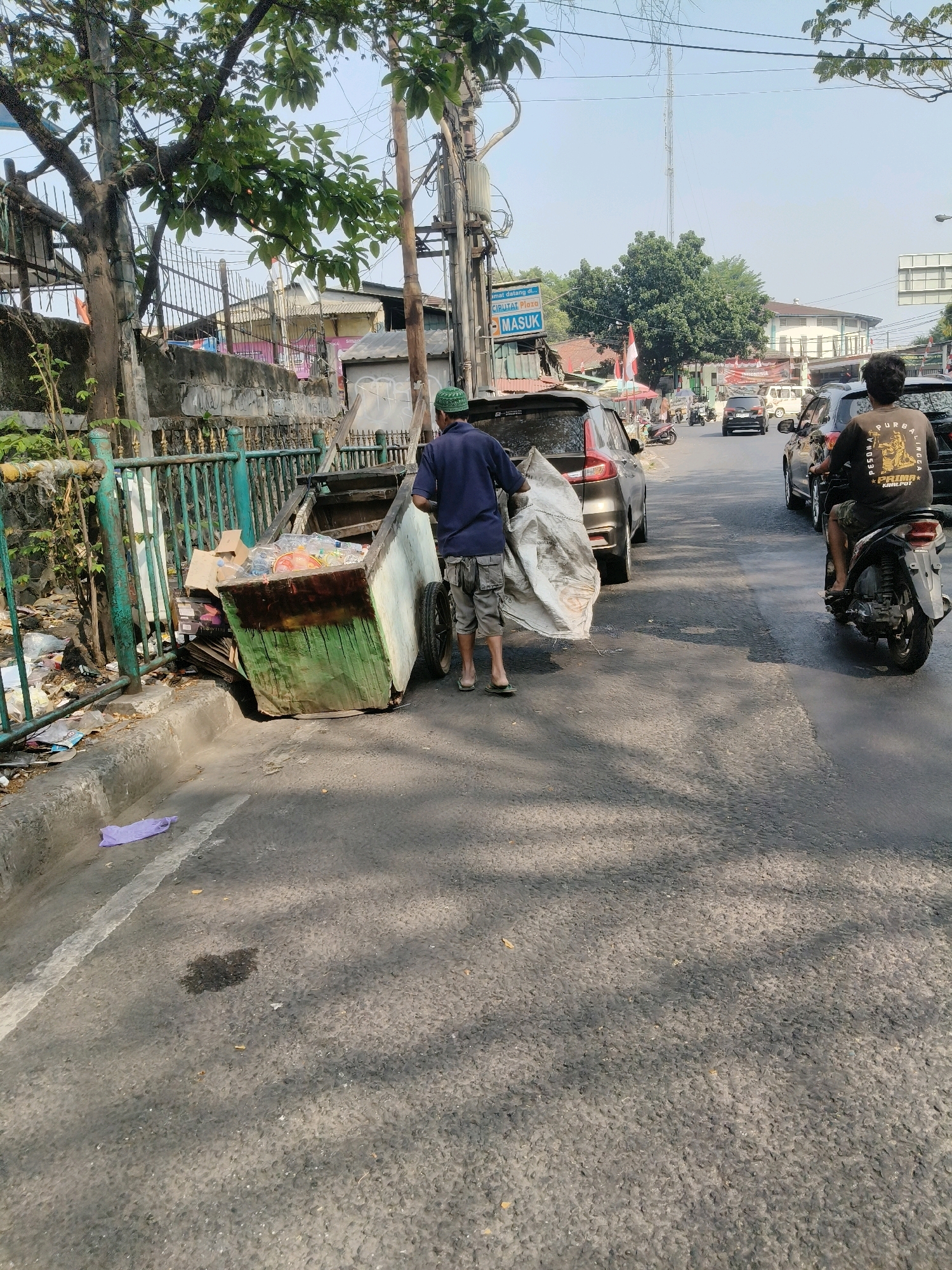 In South Tangerang, Indonesia, a man in a blue shirt and patterned shorts is pushing a cart filled with trash along the side of a street. The cart, which appears to be made of wood and metal with a green painted section, is heavily laden with various discarded items, including plastic bottles and bags. Adjacent to the cart, on the sidewalk, is more accumulated trash, some of which has spilled onto the curb. A large white plastic bag is being carried by the man. Several vehicles are visible on the road, including cars and a motorcycle with a rider wearing a dark t-shirt. The environment appears to be an urban setting with trees, buildings, and electrical poles lining the street. The sky is clear and bright.