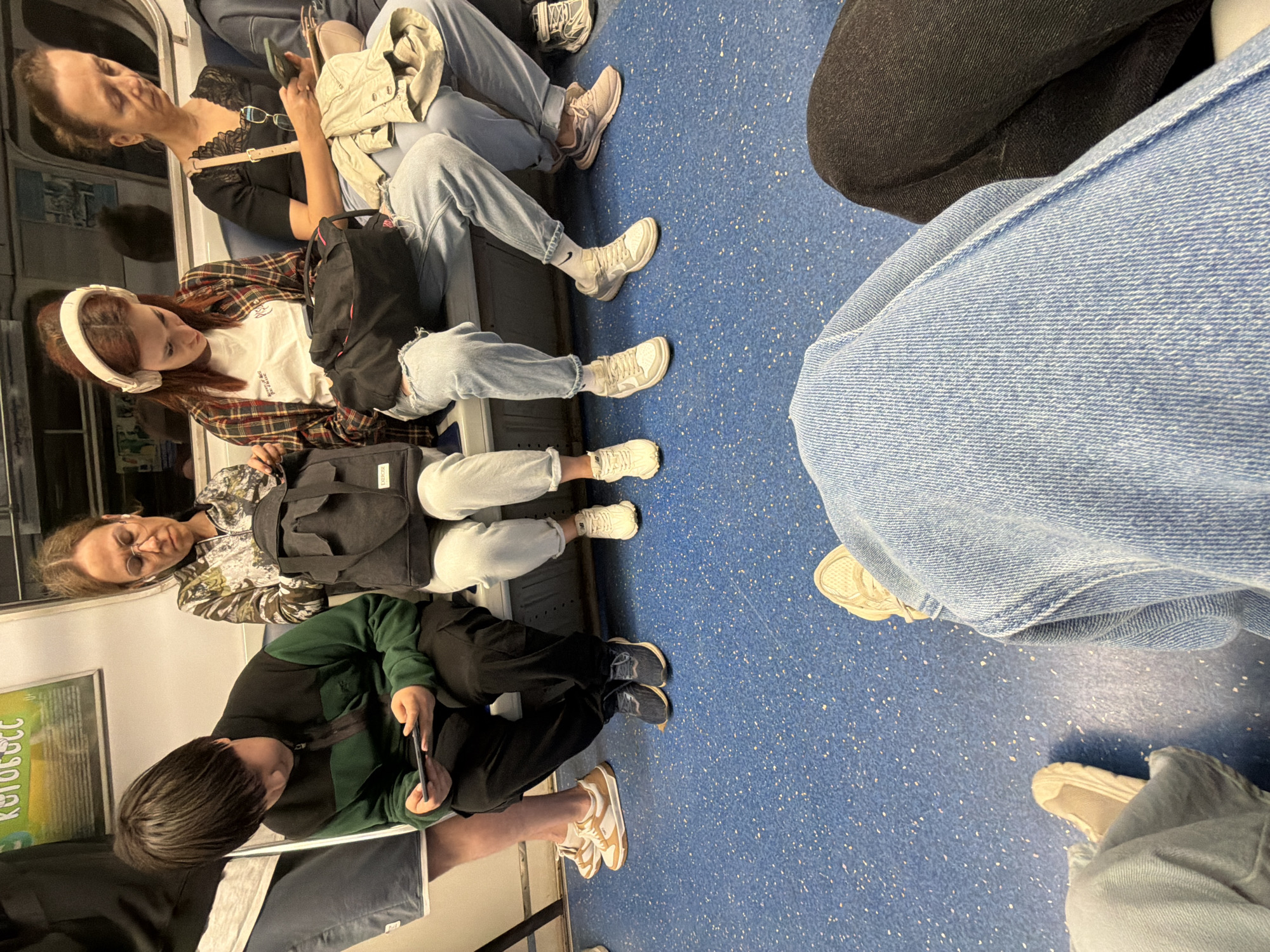 The image depicts an interior view of a subway car. Four individuals are seated on a blue bench. On the far left, a young person wearing a green and black jacket looks down at a mobile device in their hands. Next to them, a woman in a camouflage patterned jacket with glasses is looking down. To her right, a woman with headphones on and wearing a plaid shirt is also looking down. On the far right, a woman in a black top and light blue jeans is holding a mobile phone. The floor of the subway car is blue with white speckles. The overall atmosphere is one of quiet transit, with each person engaged in their own activity.