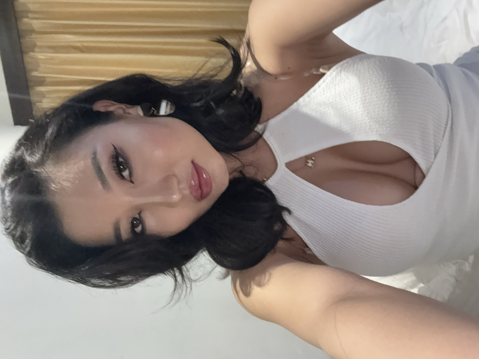 A close-up, portrait-style photo captures a woman indoors in North Kuta, Indonesia, looking directly at the viewer. She is positioned on her side, with her head tilted, and her body slightly reclined, suggesting she might be lying on a bed or a similar surface.
The woman has dark, wavy hair, styled loosely around her face and shoulders. Her skin appears fair with some blush on her cheeks. She has prominent eye makeup, including winged eyeliner and defined brows, and her dark brown eyes are open. Her lips are full and glossy, with a reddish-pink tint, slightly parted. She wears small, white and striped earrings that are partially visible in her left ear.
She is dressed in a fitted, white ribbed top with a cut-out detail on the chest, revealing cleavage. A delicate gold-colored necklace with a small pendant is visible around her neck. Her left arm is slightly bent, and her right arm is also visible, resting near her head.
The background consists of horizontal, golden-toned blinds or curtains on the left side, and a light-colored, possibly white, sheet or bedding on the right side. The lighting appears soft and even, without strong shadows, suggesting an indoor setting with either indirect natural light or artificial illumination. There is no visible text in the image.
