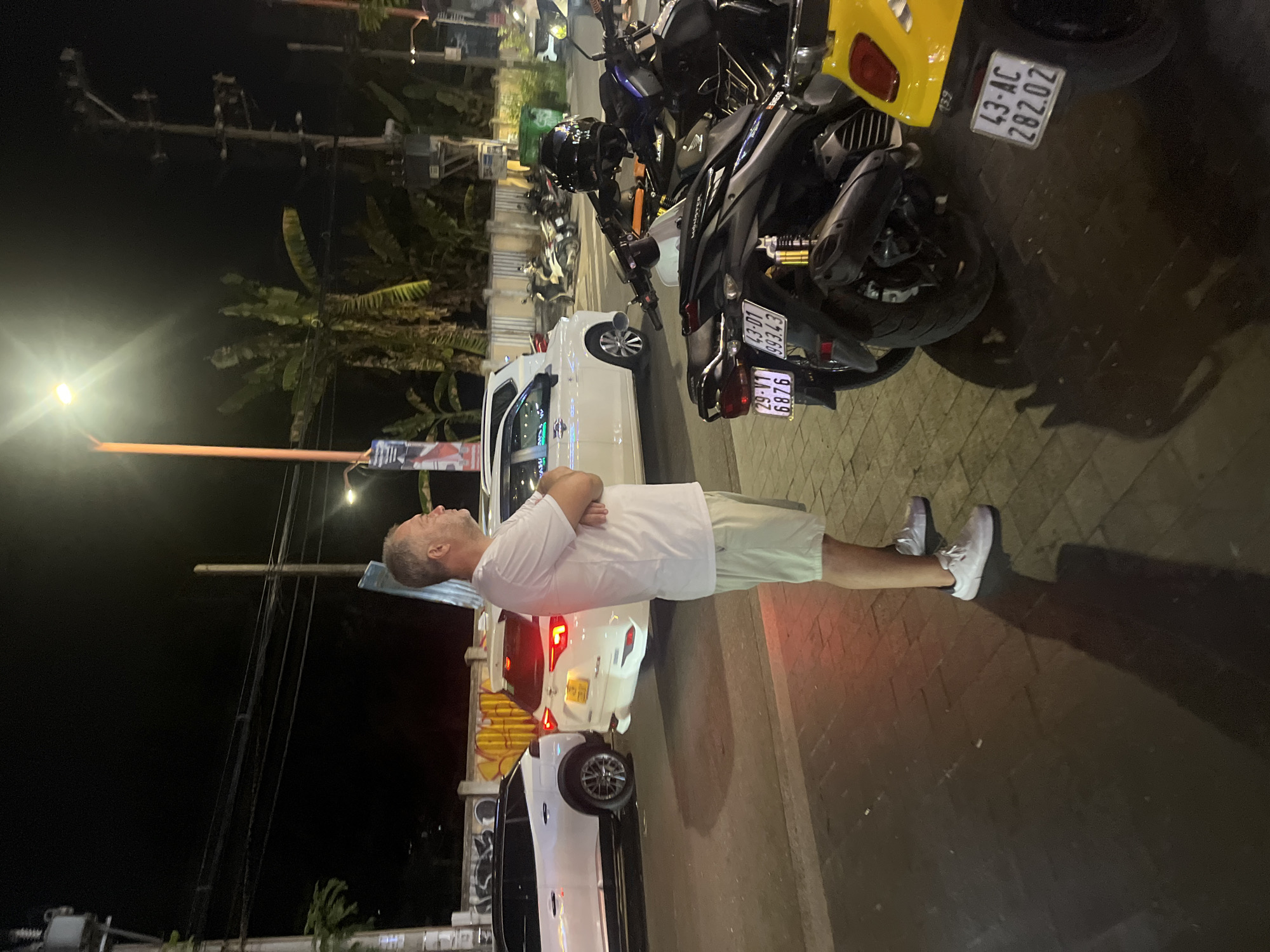 A man stands on a sidewalk at night, with his arms crossed and looking up. He is wearing a white t-shirt and light-colored shorts with white sneakers. The scene appears to be in a city, possibly Đà Nẵng, Vietnam, given the context. Behind him, several motorcycles are parked. One motorcycle is yellow, and another is black with racing-style decals. A white van is also parked nearby. The street is paved with brick-like stones, and there are overhead power lines and streetlights. The time of day is night, and the lighting suggests it is a clear night. The overall mood is casual and relaxed.
Visible text includes license plates on the motorcycles. One license plate reads "43-AC 282.02". Another reads "29-Y1 6876". There are also some stickers on the motorcycles, but the text is not legible. There is some graffiti visible on a structure in the background.