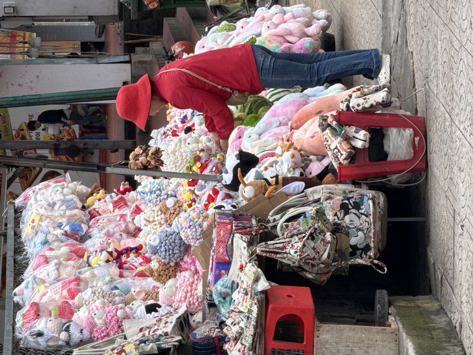 An outdoor market stall in Da Lat, Vietnam, displays a large assortment of plush toys, primarily rabbits and sheep, in various colors and sizes. The toys are arranged on racks and in baskets, some still in transparent plastic packaging. A woman wearing a red hat and a red jacket, along with blue jeans and white shoes, is interacting with the merchandise, appearing to be a vendor or a customer. In the background, a man in a dark shirt and grey pants is visible, and further back, a sign with Vietnamese text is mounted on a building. The ground is paved with textured tiles, and the overall atmosphere suggests a commercial street scene.