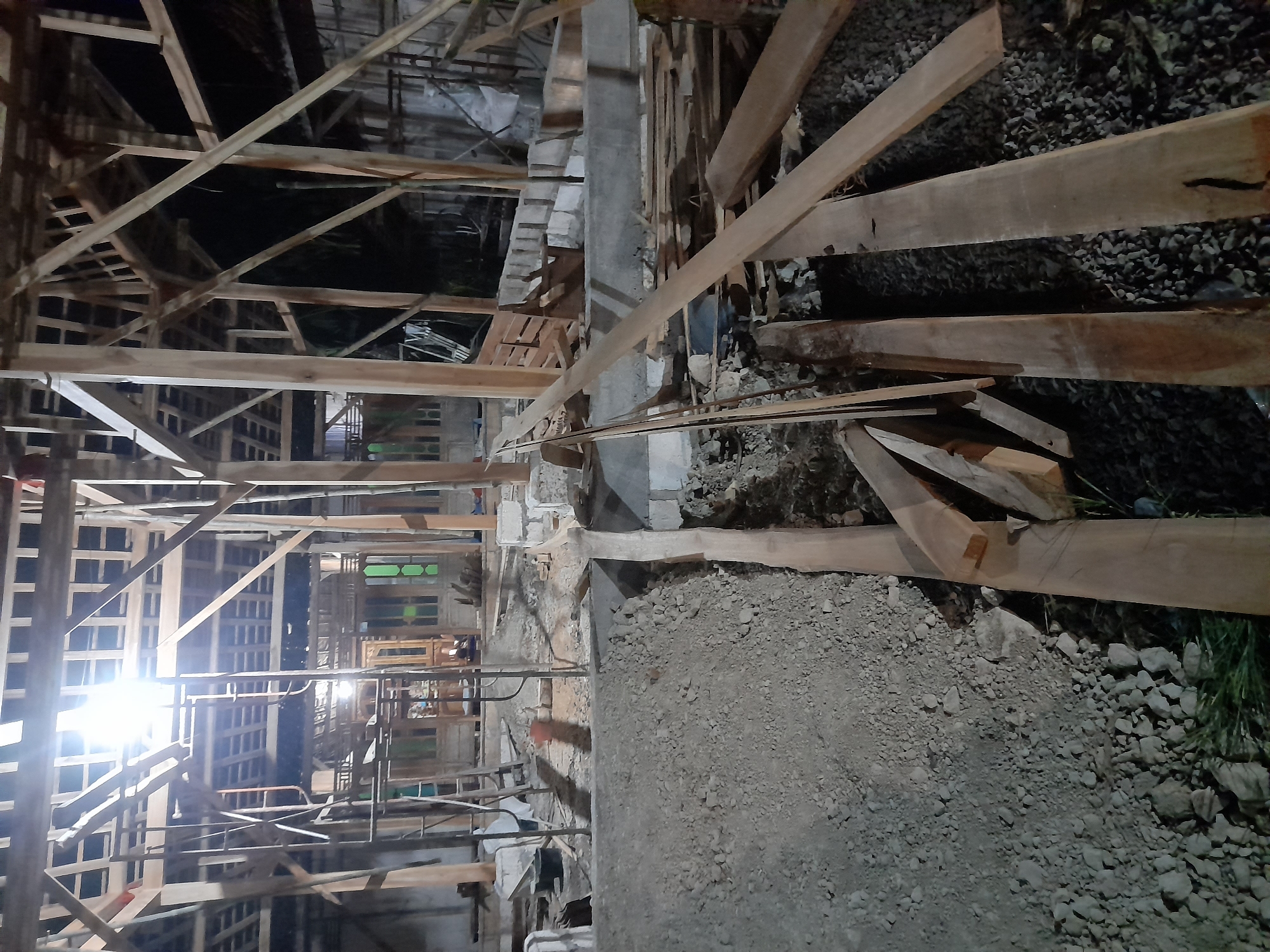The image depicts an outdoor construction site in Pulokulon, Indonesia, during nighttime. The primary subject is a partially constructed wooden building, with exposed wooden beams and supports forming the framework of the roof and walls. Scaffolding is visible, indicating ongoing work. The ground is covered with gravel and dirt, with piles of construction materials, including lumber and concrete blocks, scattered throughout the scene. A bright overhead light illuminates the area, casting shadows and highlighting the unfinished structure. There are no people visible in the image.
