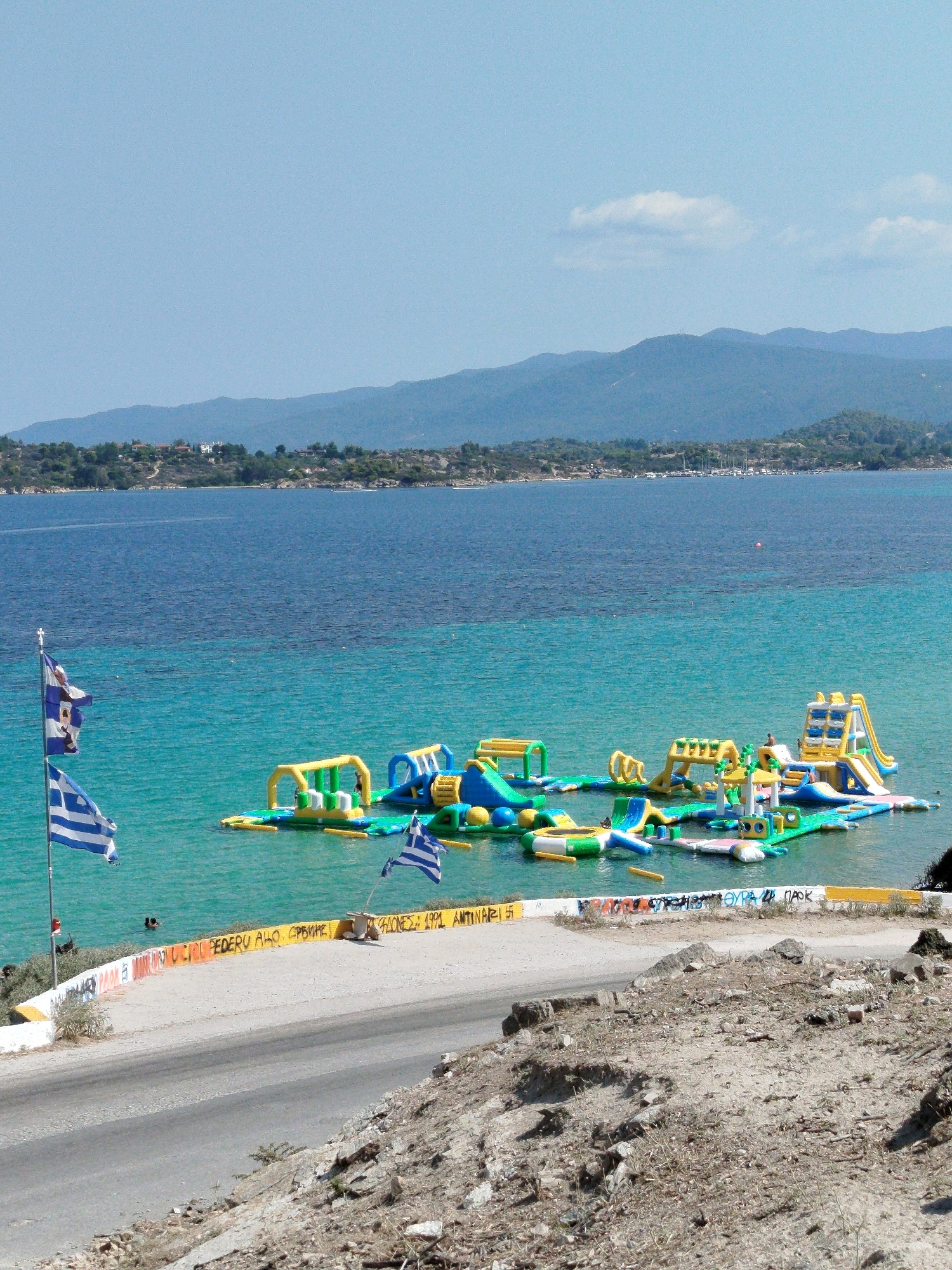 The image captures a sunny day at a coastal location in the Sithonia Municipal Unit, Greece. In the foreground, a paved road runs alongside a rocky, uneven embankment dotted with sparse vegetation. Beyond the embankment, a retaining wall is covered in graffiti and yellow warning paint. Two Greek flags are prominently displayed on flagpoles on the left side of the scene, one flying slightly above the other.
The middle ground features a body of bright turquoise water where a large inflatable water park is set up. The water park consists of numerous interconnected inflatable structures, including slides, climbing walls, trampolines, and obstacles, in vibrant yellow, blue, and green colors. A few people can be seen on the inflatable structures, enjoying the recreational activities. Further out in the water, a small cluster of boats is visible.
The background is dominated by the deep blue of the sea, transitioning to a hazy horizon where a range of mountains forms the coastline. The mountains are covered in lush green vegetation, with scattered buildings visible on their slopes, suggesting a town or village. The sky above is a clear, light blue with a few wispy white clouds. The overall scene evokes a sense of summer vacation and outdoor recreation.