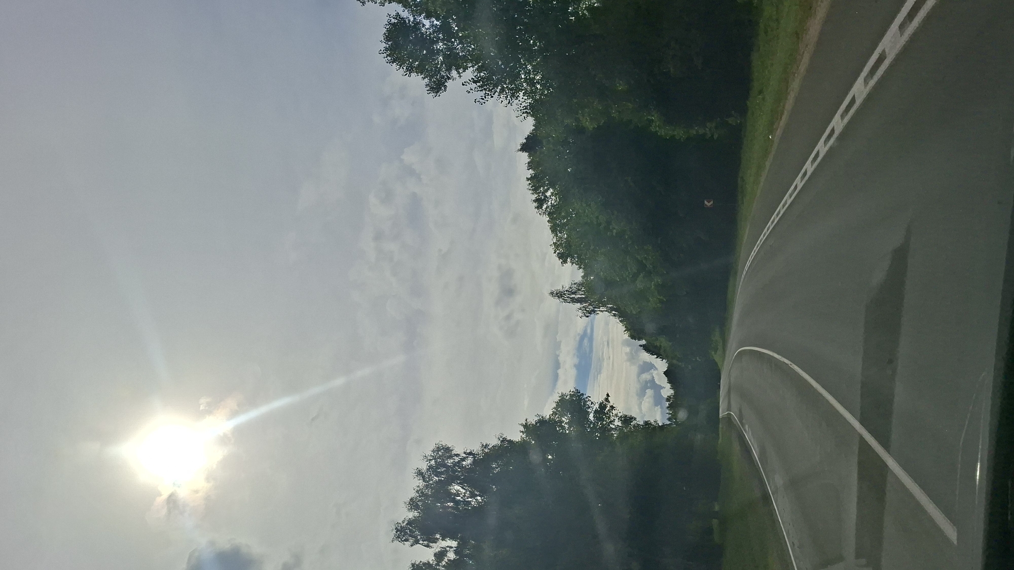 The image depicts a view from a vehicle traveling on a paved road that curves to the left, flanked by dense green trees on both sides. The sky is overcast with bright sunlight breaking through the clouds on the left side, casting a bright glare. The road has white lane markings, including a solid white line on the left and a dashed white line on the right. A small orange directional sign is visible on the right side of the road. The environment appears to be rural, possibly a highway or a well-maintained country road, consistent with the location of Мядзельскі сельскі Савет, Belarus, known for its natural landscapes. There are no people or animals visible in the scene. The time of day appears to be daytime, given the presence of sunlight.