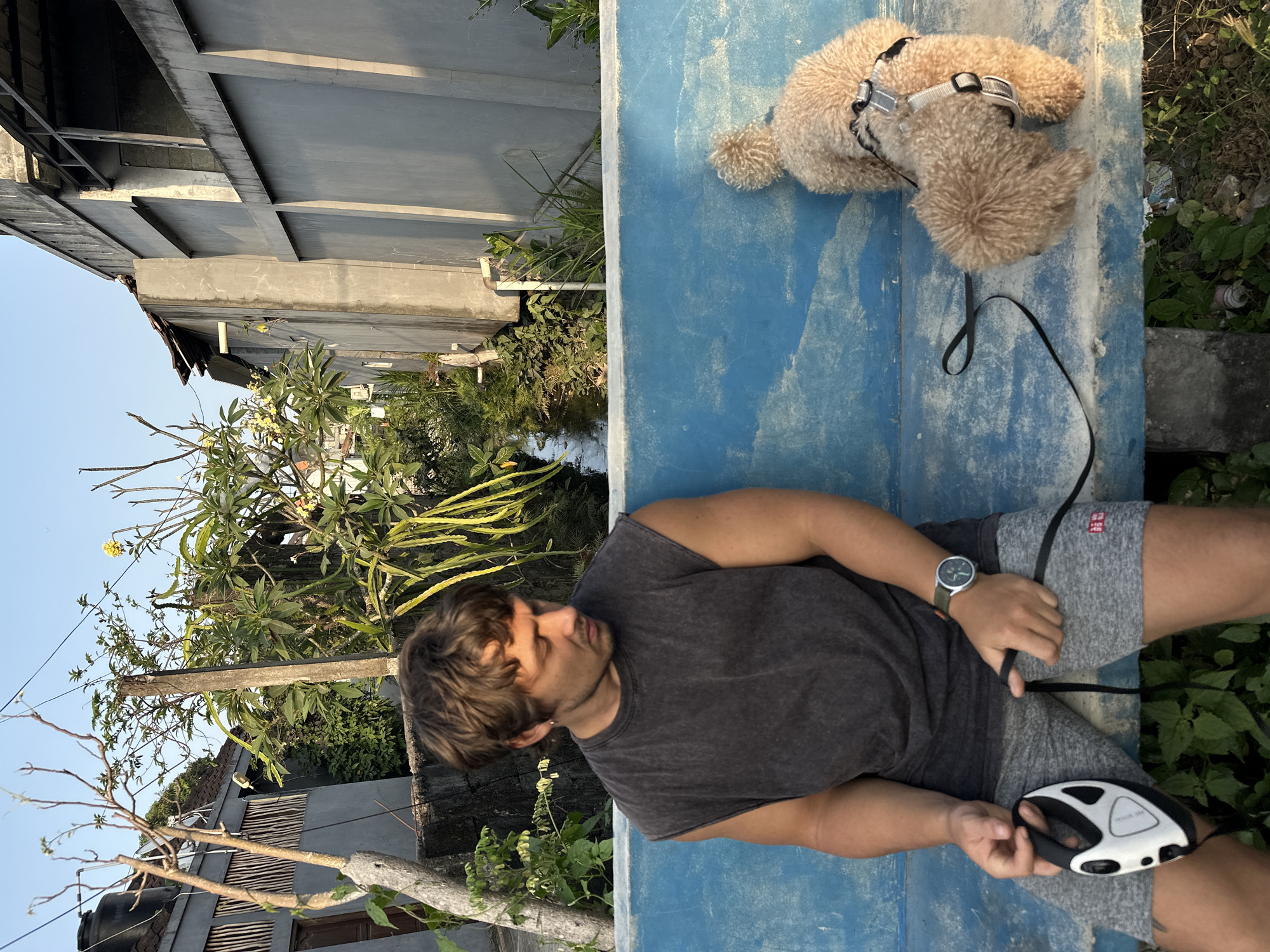 In North Kuta, Indonesia, a young man with brown hair and a dark gray, sleeveless t-shirt sits on a vibrant blue bench. He is looking down, holding a black and white retractable dog leash in his left hand and the leash itself in his right. His gray shorts have a small red logo. A light brown poodle, wearing a gray harness and black leash, stands on the bench to his right, facing away from the camera. Behind them, a narrow, water-filled channel runs horizontally, flanked by lush green vegetation and the walls of concrete buildings. The sky above is clear and blue.