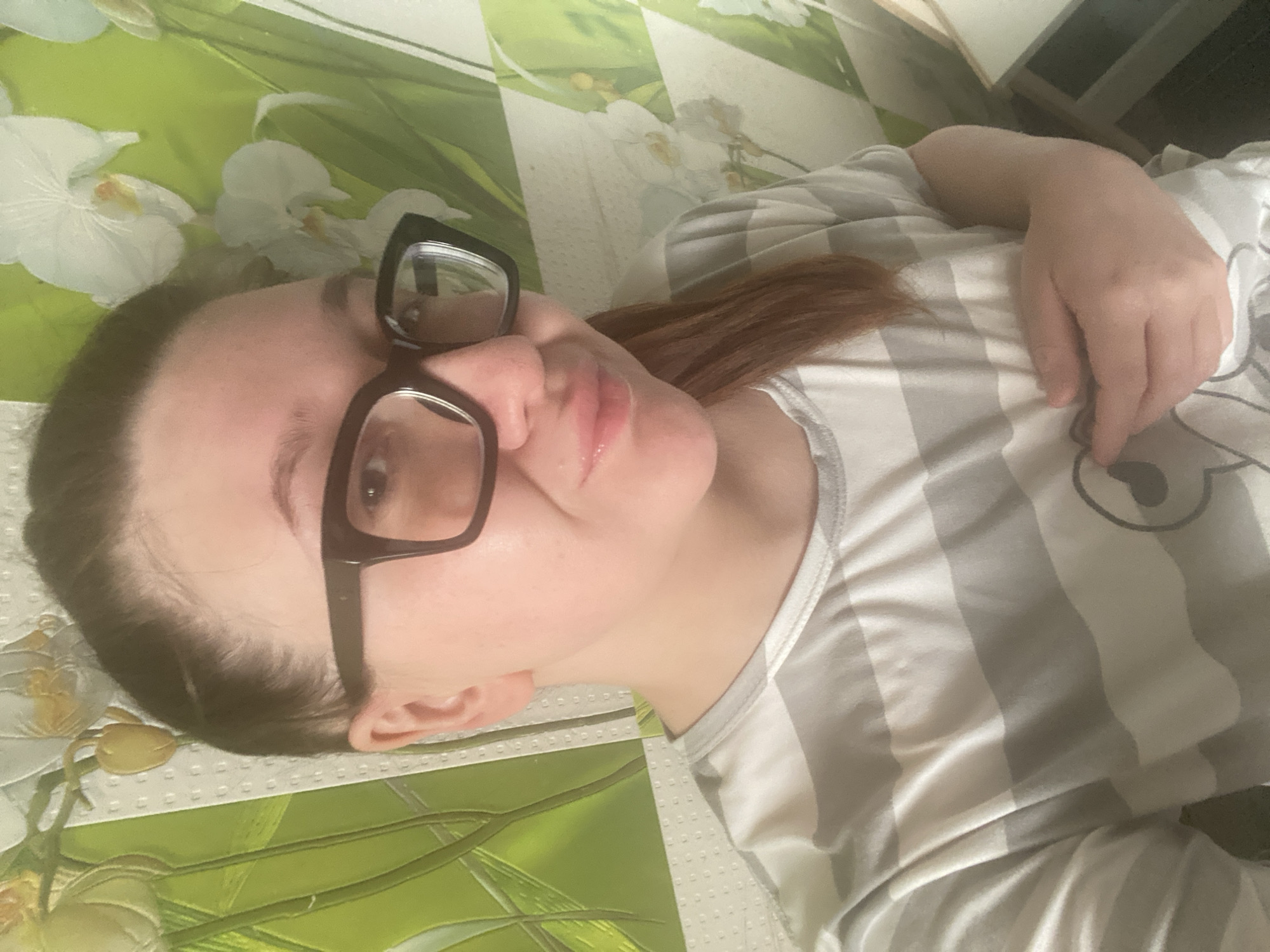 A woman with light skin, brown hair with red undertones, and black framed glasses lies on her back. She is wearing a light grey and white striped shirt. Her right hand rests on her chest. She appears to be on a bed with a green and white floral patterned bedspread. The location is unknown and there is nothing to suggest it is in Chaikovsky, Russia.