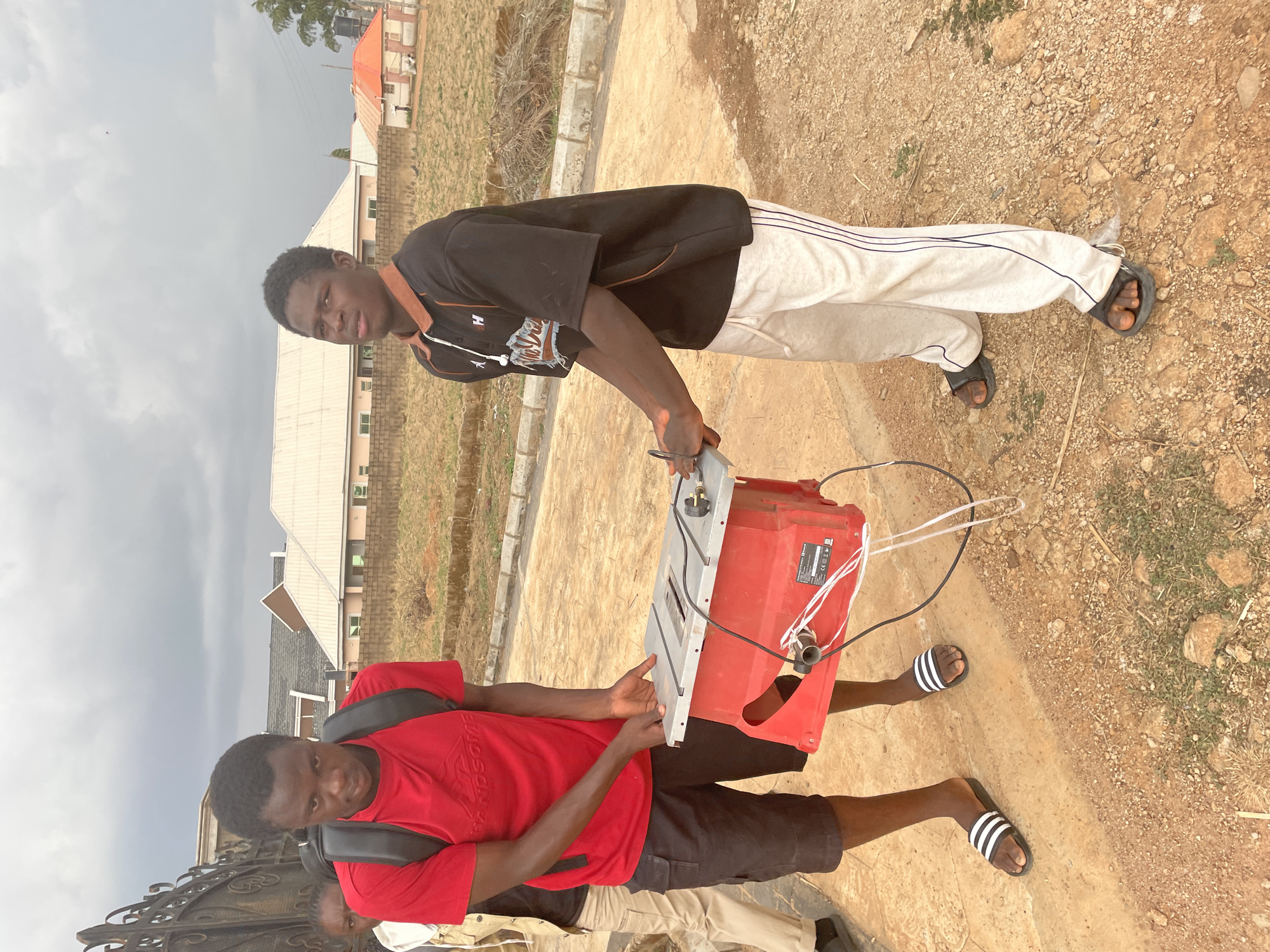 Two young men are outside. One man, wearing a red t-shirt and black shorts, is holding one end of a red power tool. The other man, wearing a black shirt and white sweatpants, is holding the other end of the tool. They are standing in a dusty area with some grass and sparse vegetation. In the background, there are buildings with pitched roofs. Given the available information, it is impossible to confirm the specific location within the city of Jos, Nigeria.