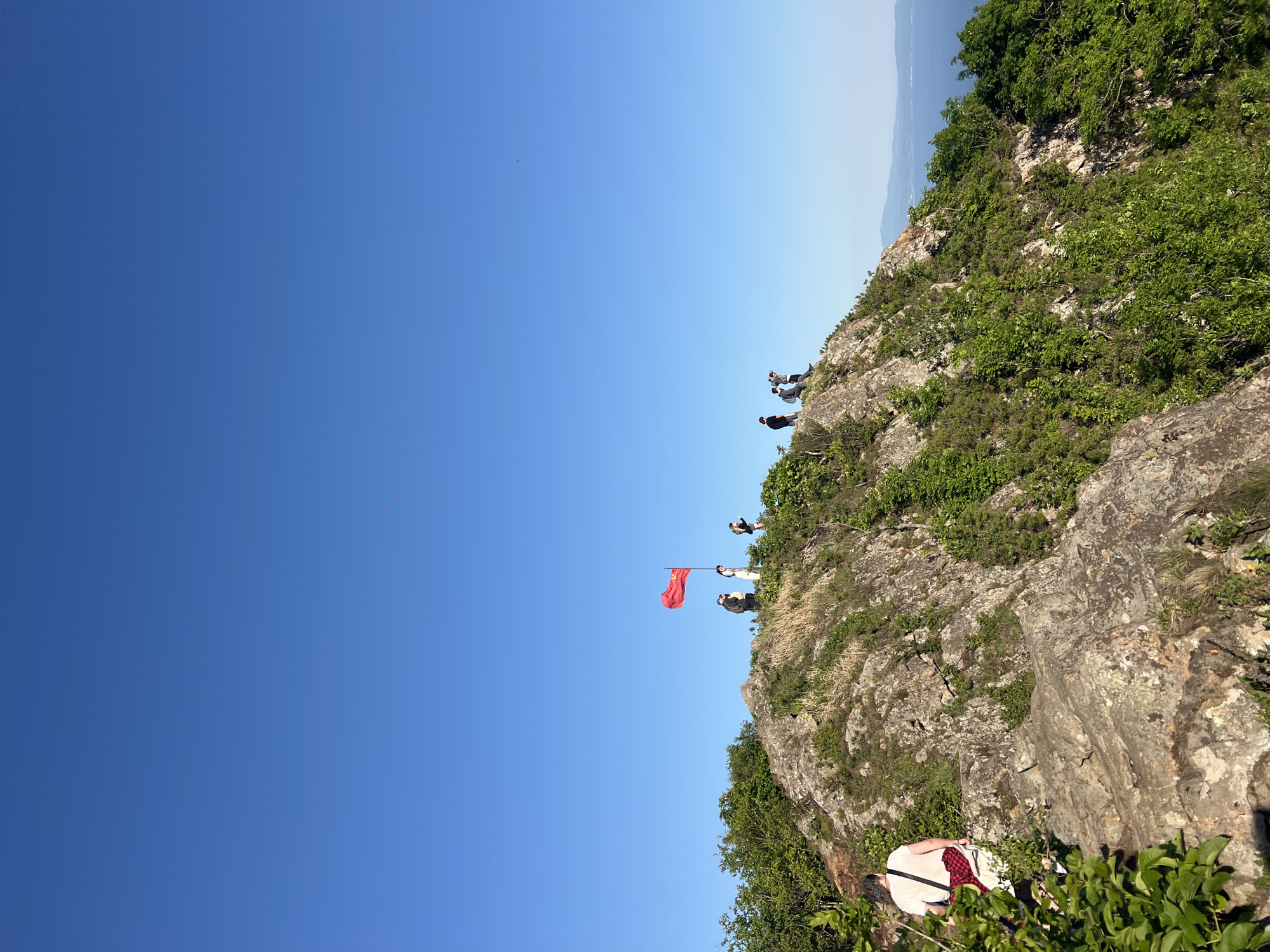The image depicts a rocky cliff with sparse vegetation under a clear blue sky, likely on a sunny day. In the background, hints of a distant, hazy landscape are visible. Several individuals are positioned on the cliff's edge; at least four people are standing near a red flag. A person in a white shirt and a red-and-black checkered pattern is partially visible in the lower part of the image. The cliff's surface consists of rock and patches of greenery. The location is likely Vladivostok, Russia.