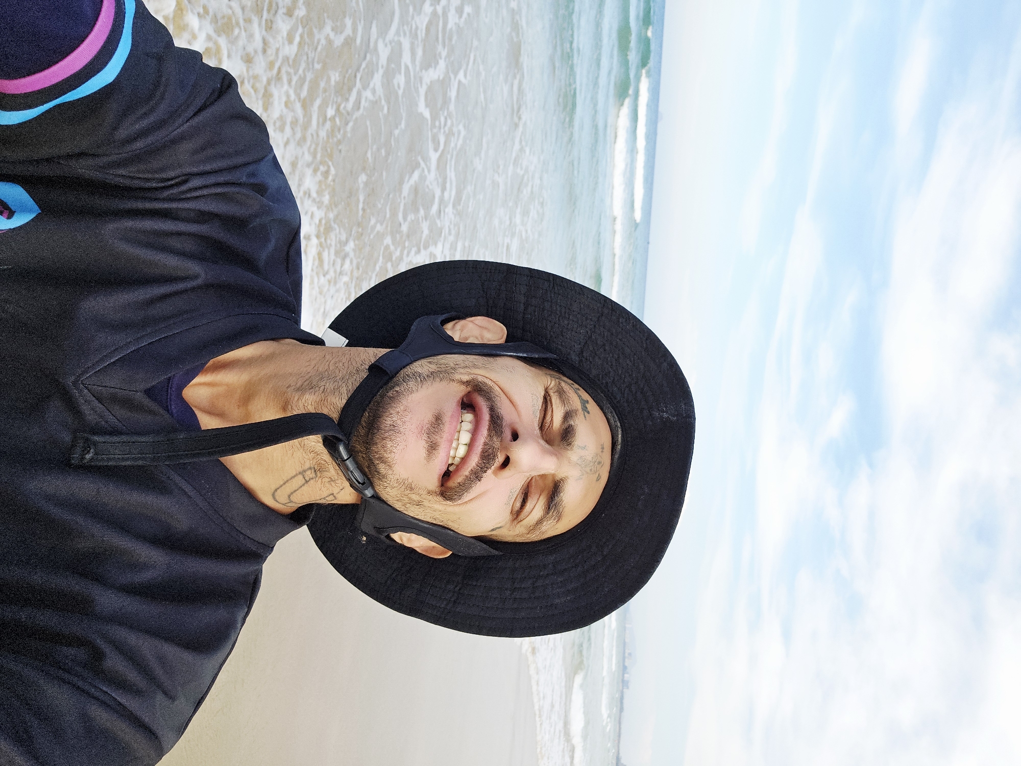 A selfie, rotated clockwise, shows a man smiling at the camera. The man is wearing a black bucket hat with a strap, and a black long-sleeved shirt with blue and pink accents on the sleeve. He has tattoos on his face and neck. The background shows a beach with white sand, ocean waves, and a light blue sky with some clouds. The location is likely Cam Lâm, Vietnam, based on the prompt.