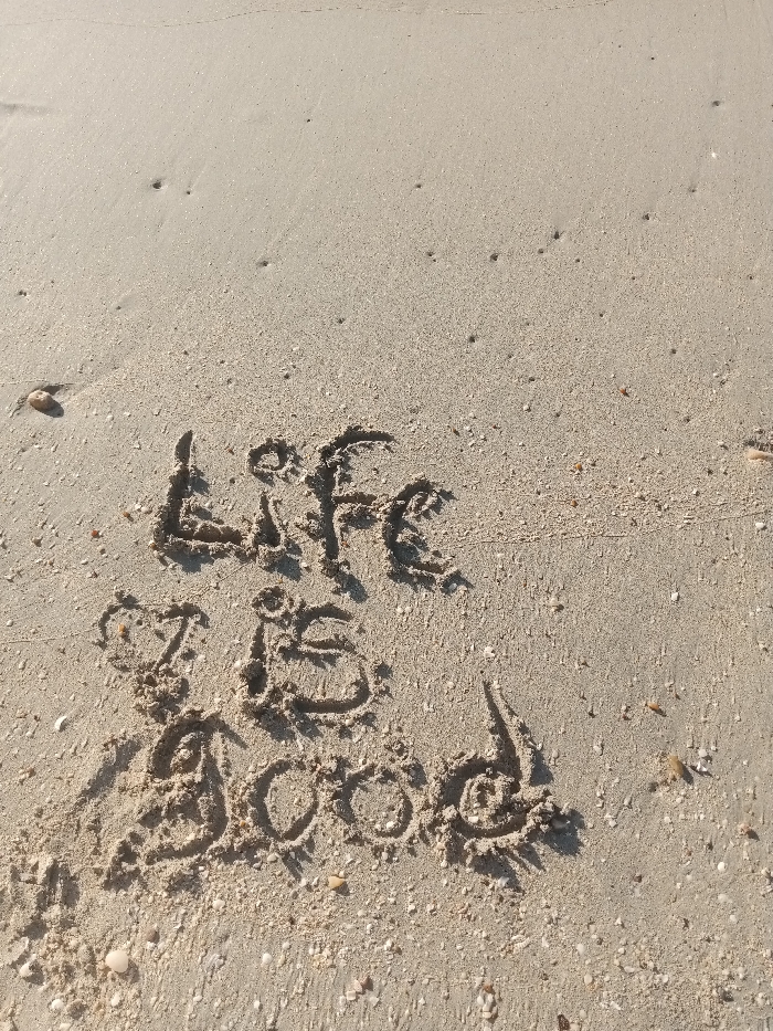 The phrase "Life is good" is written in the sand. The letters are formed by displacing the grains of sand, creating a darker contrast against the surrounding lighter sand. Small shells and pebbles are scattered around the inscription. The sand appears smooth and fine-grained, with some minor disturbances and ripples on its surface.