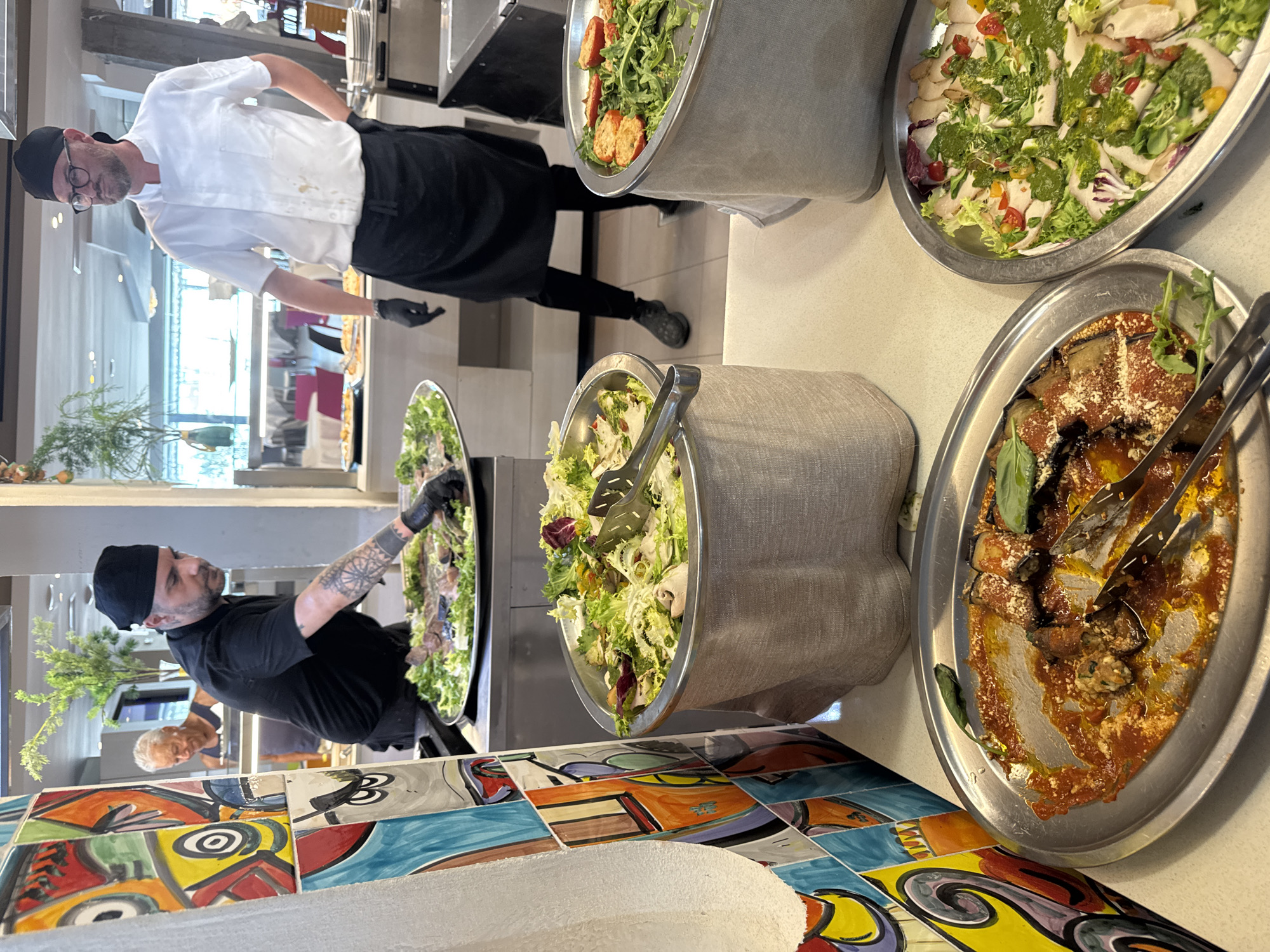This media file captures a scene at a food service counter, likely in a restaurant or catering event. Two individuals, both men, are present. One man, wearing a white chef's coat, black pants, a black skullcap, and glasses, appears to be serving or arranging food from a large metal tray. He is wearing black gloves. His posture suggests he is actively engaged in his task. Another man, wearing a black t-shirt and a black skullcap, is also visible in the background, seemingly observing or assisting. He has tattoos on his left arm and is also wearing black gloves, reaching into another large metal tray filled with food.
Several large metal serving dishes are prominent. One contains a salad with various greens, tomatoes, and a green dressing. Another dish holds a vegetable-based dish with red sauce and cheese. A third, larger metal container, appears to hold a salad with mixed greens and vegetables. The food items suggest a buffet or self-service setup.
The background shows a bright, indoor setting with reflections on glass surfaces, indicating a well-lit environment. The foreground features a colorful, tiled surface with an abstract, artistic design, adding a decorative element to the scene. The overall atmosphere seems professional and focused on food preparation and service. No specific time of day or weather conditions are discernible. The location context provided suggests the scene is set in Isola delle Femmine, Italy.