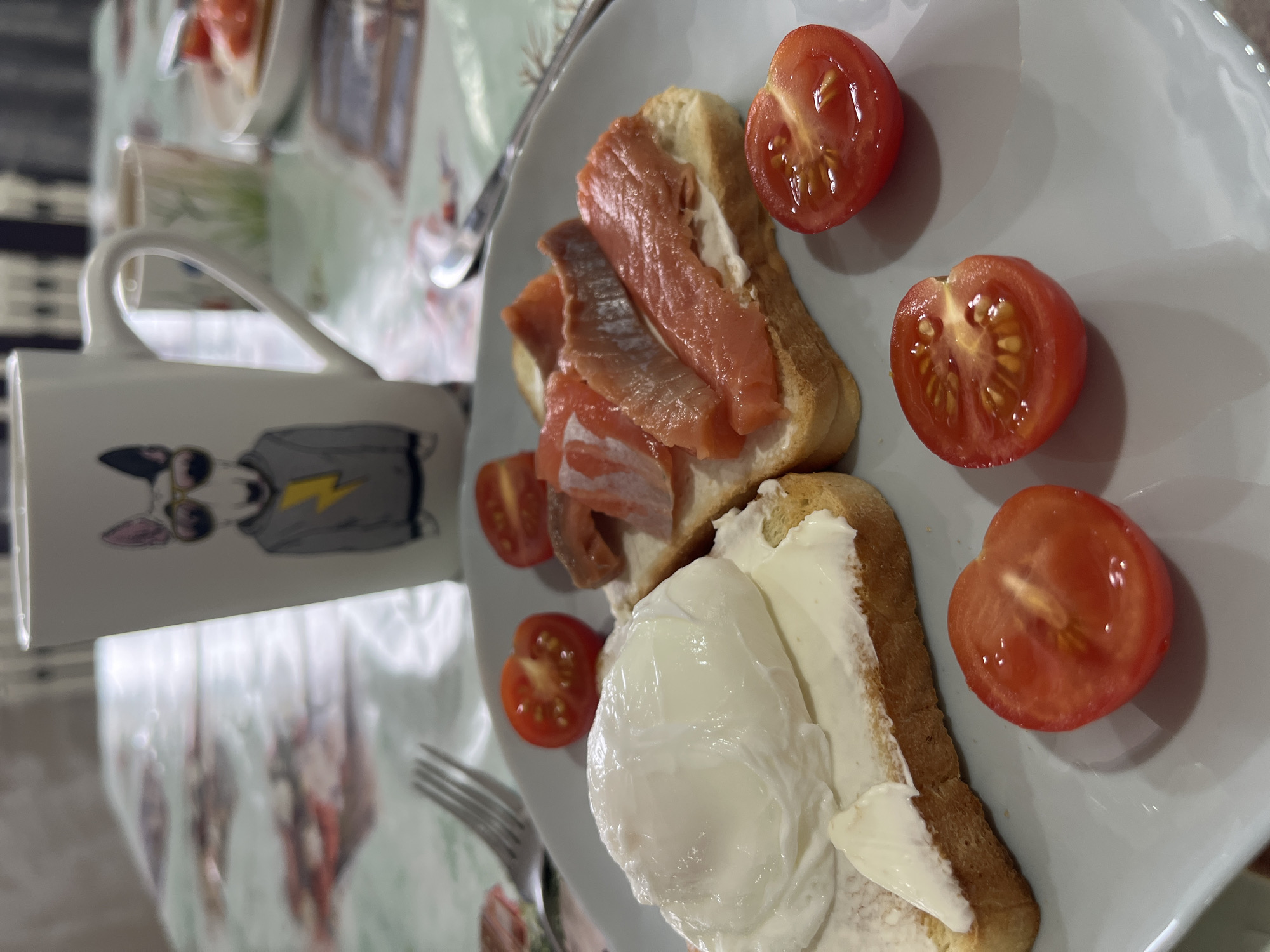 A plate contains two open-faced sandwiches: one topped with sliced salmon and the other with a poached egg and a creamy spread. Halved cherry tomatoes are scattered around the sandwiches on the plate. A mug with a cartoon graphic is partially visible, as are a fork and a spoon. The location is Chaikovsky, Russia.