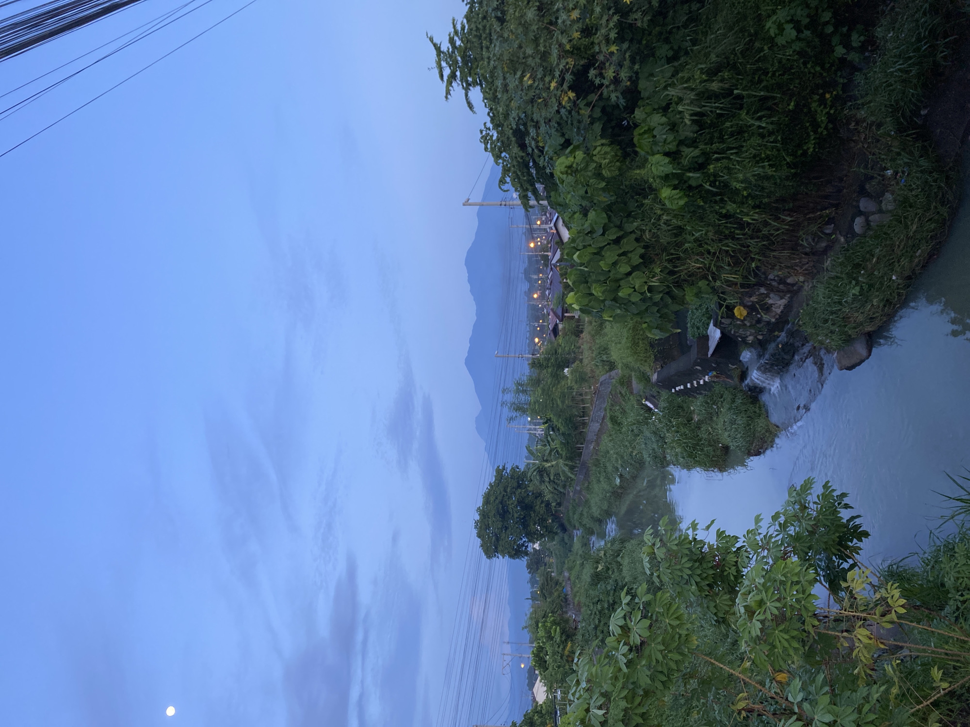 The image, oriented horizontally, depicts a tranquil natural landscape transitioning from day to night, likely captured at dusk or dawn in Davao City, Philippines. The upper portion of the scene is dominated by a clear, soft blue sky with wispy, scattered clouds. A distinct, bright moon is visible in the lower left area of the sky. Multiple black utility lines stretch diagonally across the top left corner of the sky, indicating overhead power or communication infrastructure.
In the mid-ground and extending into the foreground, a winding river or stream flows from the right side of the frame towards the left. The water appears calm and reflective in parts, with some gentle cascades or rapids visible where the terrain drops. Both banks of the river are densely covered with lush, vibrant green vegetation, including various types of trees, bushes, and tall grasses. Rocks are visible along the water's edge in several places.
In the background, a range of prominent mountains or large hills rises majestically against the horizon, their forms subtly silhouetted against the fading light of the sky. Along the base of these mountains, a line of numerous small, glowing artificial lights can be seen, suggesting a distant settlement, town, or road. More utility poles and power lines are visible along the riverbank, leading towards these distant lights.
The time of day is clearly twilight, characterized by the soft blue sky, the visible moon, and the artificial lights beginning to illuminate the distant landscape. There are no people visible in the image, nor is there any discernible text. The scene conveys a sense of peaceful natural beauty with subtle hints of human presence in the distance.
