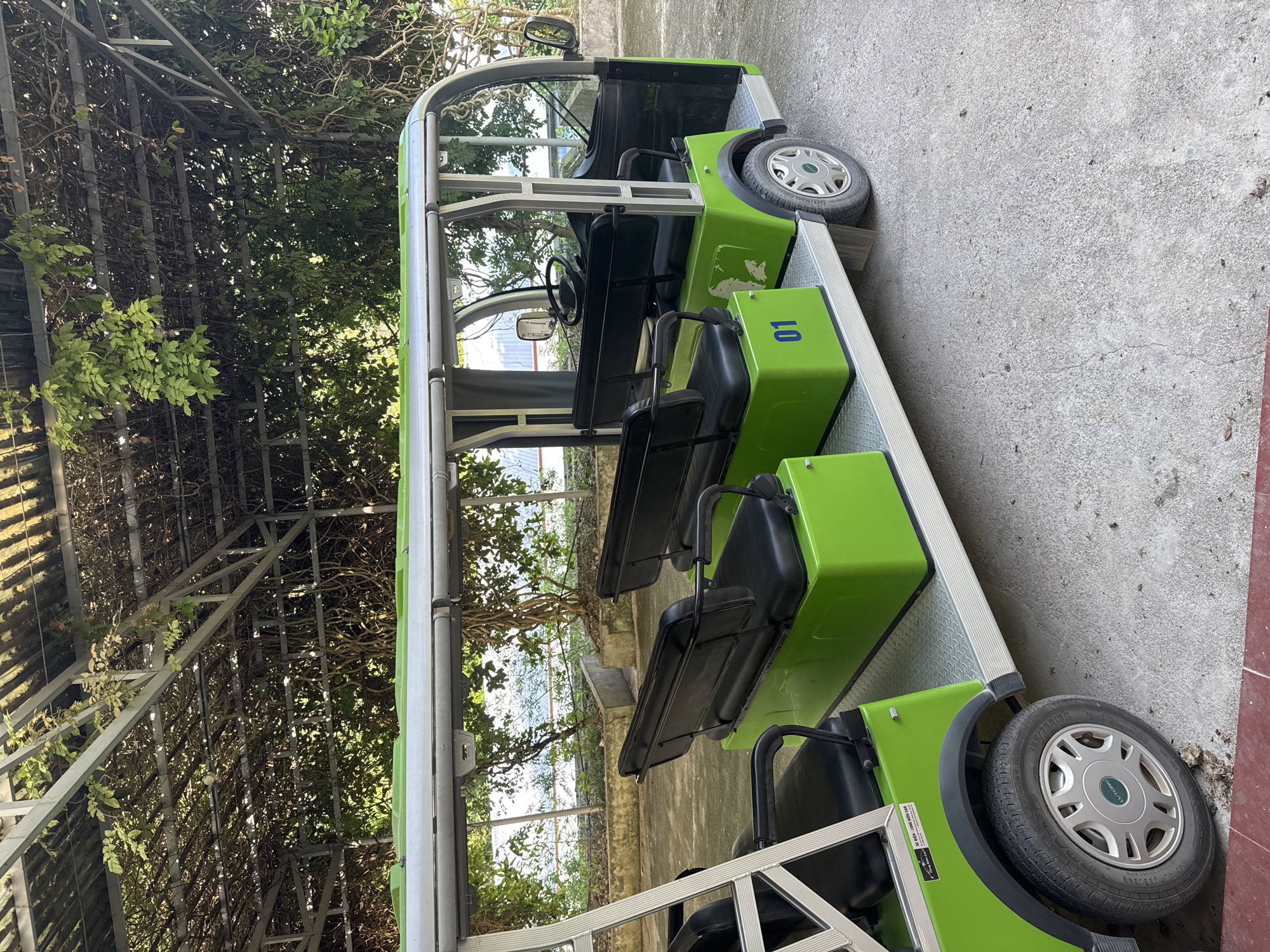 A green and silver electric cart with multiple rows of black seats is parked on a concrete surface. The cart has the number "01" in blue on the front. The environment includes trees and vines growing on a metal structure. The location is identified as Sơn Vĩ Commune, Vietnam.