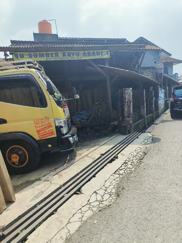 The image shows a yellow truck parked in front of a wooden building with a sign that reads "UD SUMBER KAYU ABADI 1." The building is located in Cileunyi, Indonesia, at latitude -6.947 and longitude 107.724. The truck has a sticker that reads "Sukses Itu Melewati Banyak Proses, Bukan Proses." The truck is parked on a cracked asphalt road with two drainage ditches. The truck is partially blocking the view of the building. The building is made of wood and has a metal roof. There are stacks of wooden planks in the building. There is a red brick wall behind the building. The road is lined with trees and vegetation. The sky is overcast. The scene is typical of an urban setting in Indonesia. The location appears to be a lumber yard. There is another vehicle, a dark sedan, parked in the distance to the right. The image is taken from a low angle, looking up at the truck and building. The scene is calm and tranquil, suggesting that there is no immediate danger. The image does not show any people. It is a mundane scene in the city of Cileunyi, Indonesia, at a lumber yard.