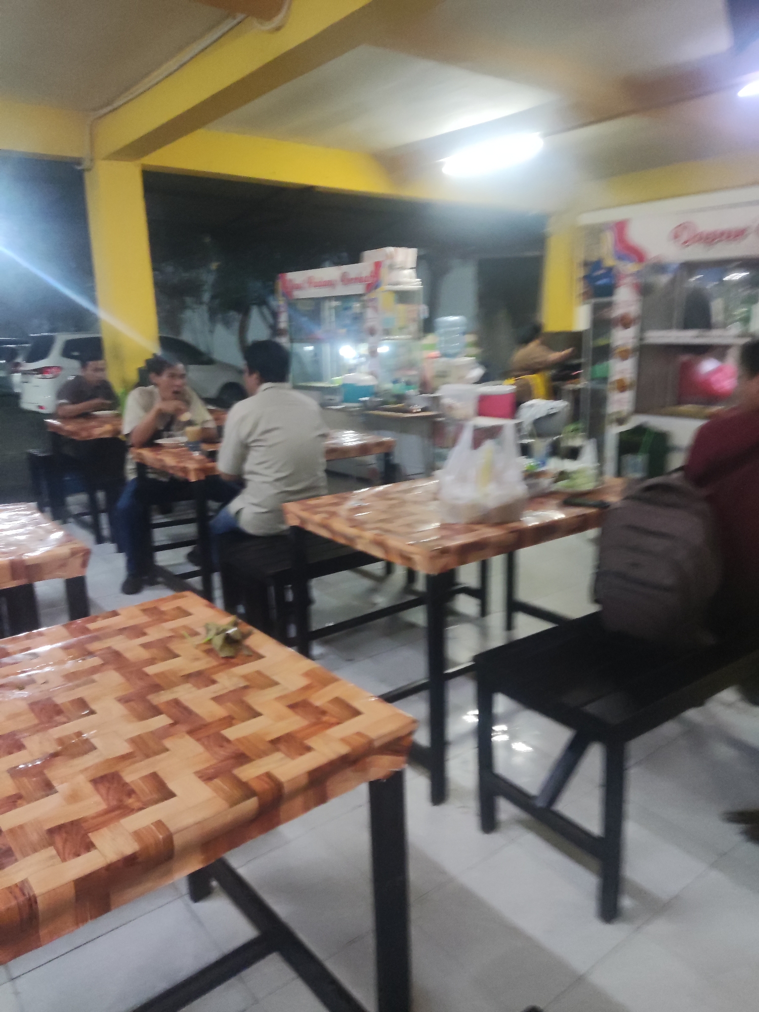 The image depicts an interior scene of an eatery in Trihanggo, Indonesia, likely at night. The establishment features several tables with distinctive patterned laminate tops, set on metal frames. Two of the tables are occupied by patrons. At a table on the left, two men are seated, one is looking at the other and holding food near his mouth. In the middle of the image, another man is seated at a table, appearing to be eating. Further to the right, a backpack rests on a dark bench next to an occupied table, suggesting another patron is present, though not fully visible. The background reveals a service counter with various items, including bottles of water and what appear to be food displays. The walls are painted yellow, and the ceiling is white with fluorescent lighting. The floor is tiled. The overall impression is of a casual dining establishment.