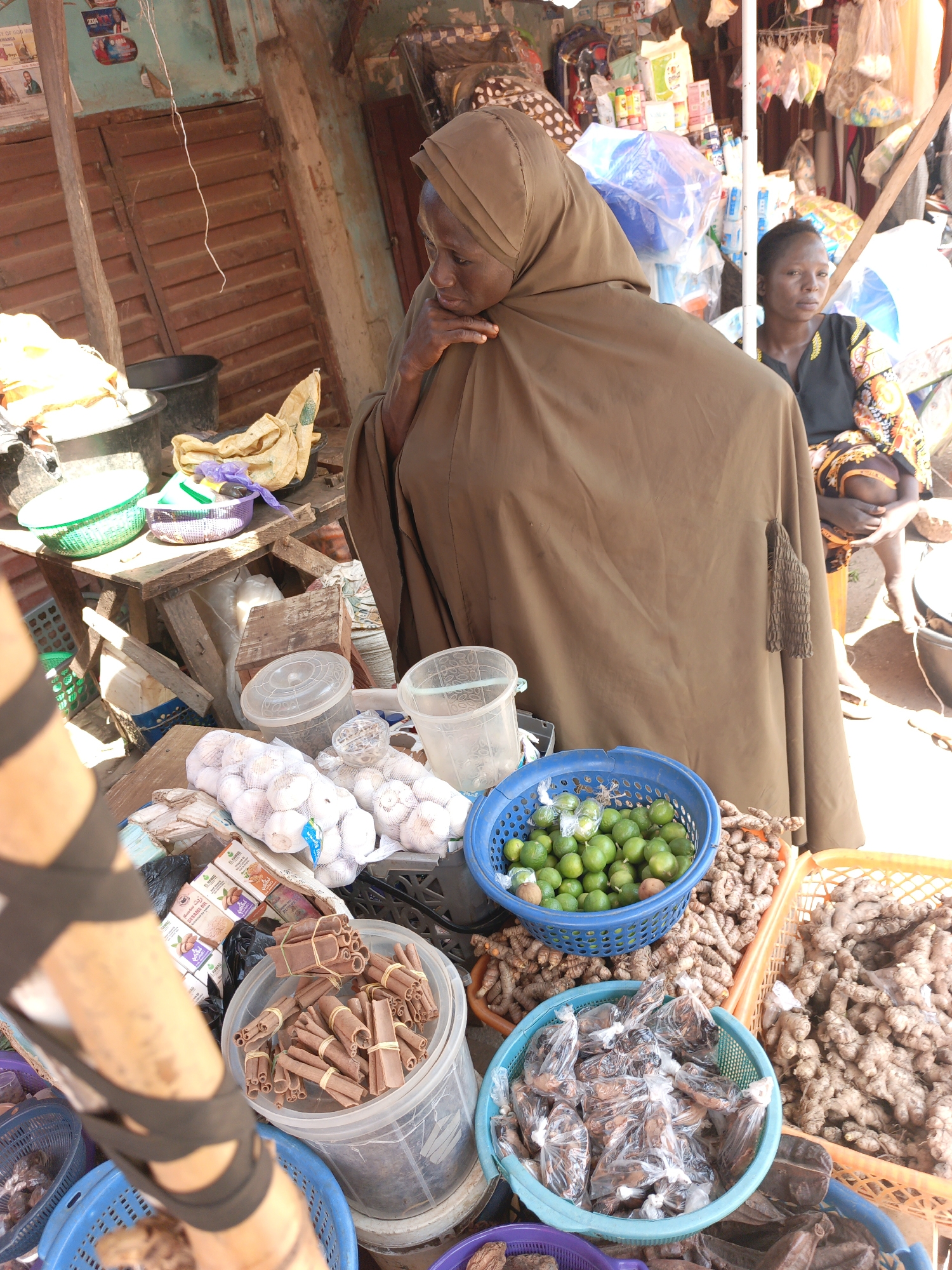 The image depicts a market scene in Akwanga, Nigeria. In the foreground are baskets and containers filled with various produce: limes, ginger, packaged goods, cinnamon sticks, and garlic. A woman wearing a brown hijab and robe is standing behind the produce, with her hand near her face as if in thought. In the background, another woman in a patterned top is seated. The market appears to be open-air with temporary structures and various goods for sale. The setting suggests a busy, everyday scene in a West African marketplace.
