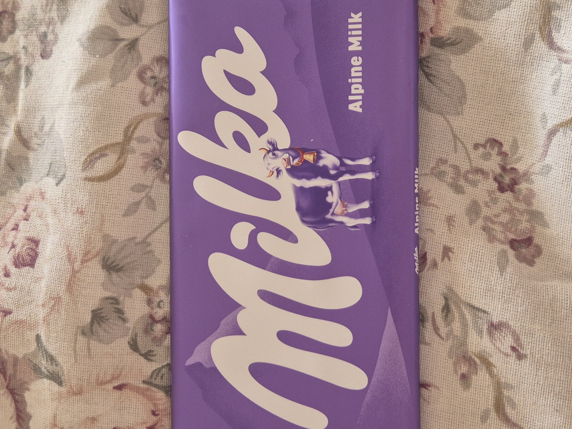 A partially visible Milka Alpine Milk chocolate bar is laying on a floral patterned fabric. The bar is purple with the white "Milka" logo and a cow illustration on the front. The text "Alpine Milk" is printed vertically in white near the edge of the bar. The location is indeterminable with the given information and no evidence to link the image to the city of Blagoevgrad, Bulgaria.