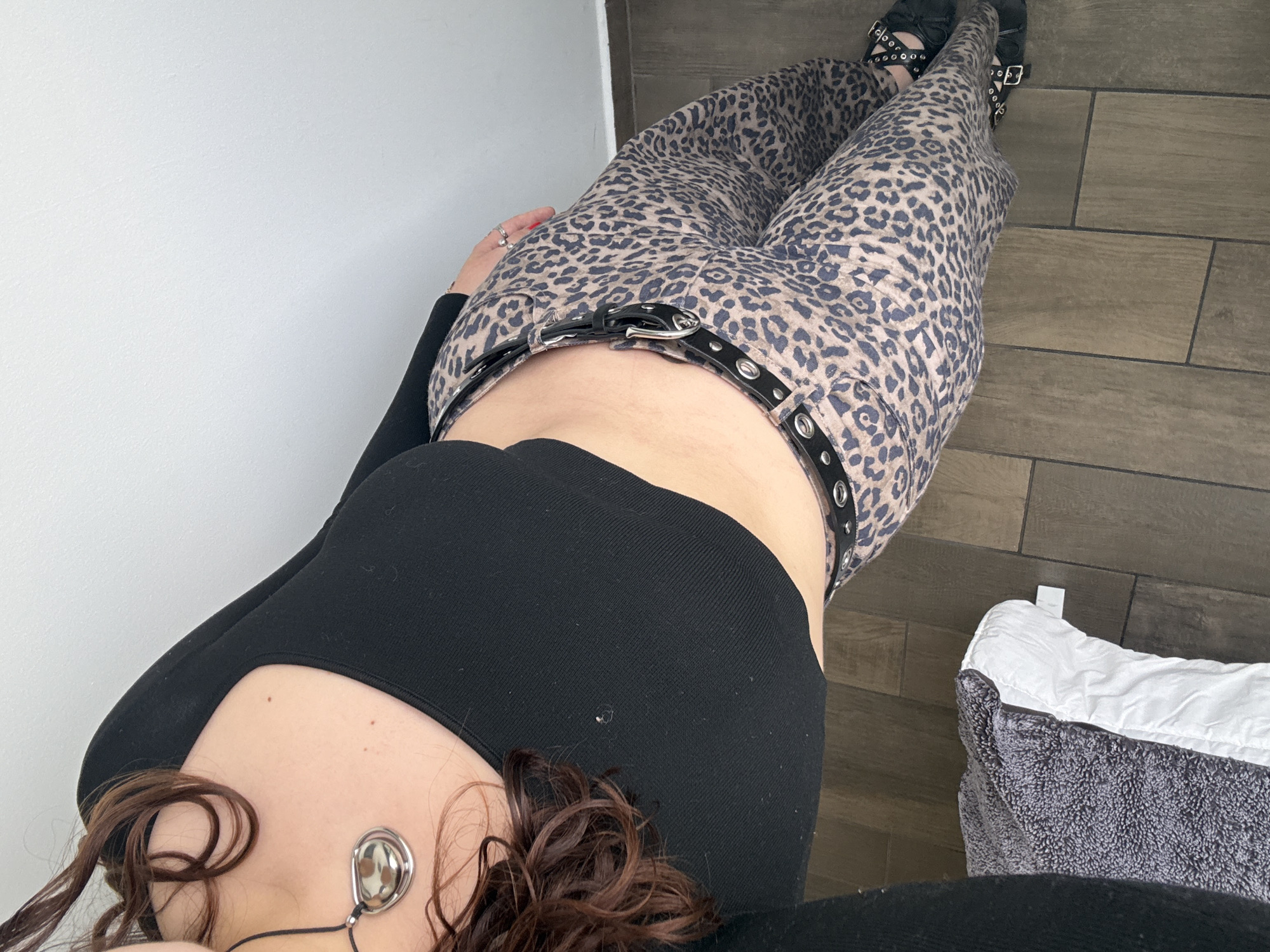 Here is a description of the image:
A full-body shot from an overhead perspective features a person wearing leopard-print pants with a black belt. The person is also wearing a black, form-fitting, long-sleeved top. Their brown curly hair is visible at the bottom of the frame, along with a necklace. The person is wearing black, studded sandals.
The background consists of a white wall, a gray tiled floor, and a gray and white folded blanket. The location of the image is unknown.