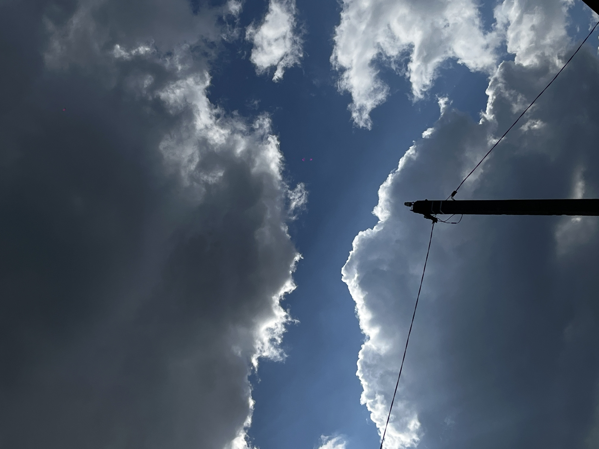 The image shows a view of the sky with clouds and a utility pole. The sky is partially clear blue, with both dark and white cumulonimbus clouds on either side. Two pink objects (possibly balloons or debris) are visible in the blue sky. A utility pole and wires extend from the bottom right corner toward the center of the image. The photograph appears to have been taken from a low angle, looking upwards. The location is assumed to be Novopokrovskaya, Russia, but there are no specific details in the image that confirm this. There are no people or actions visible.