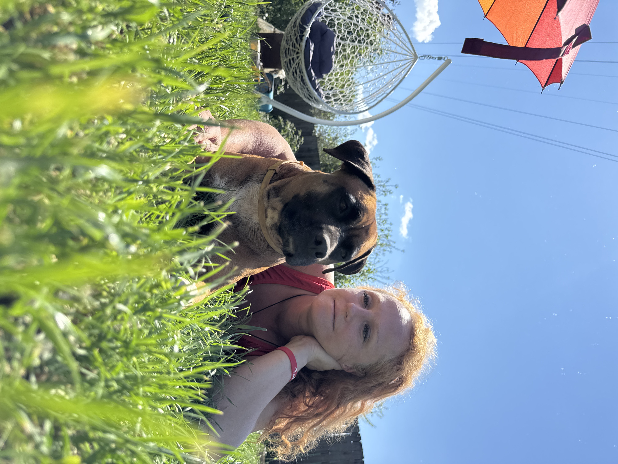 In a grassy backyard on a sunny day, a woman with long, wavy red hair lies on her side, propped up on her elbow with her hand supporting her head. Beside her, a brown dog with a black muzzle rests its head. In the background, a white hanging chair with a blue cushion is visible, along with a portion of a colorful umbrella and a wooden fence. The scene is set in the city of Chaikovsky, Russia, under a clear blue sky with some scattered clouds.