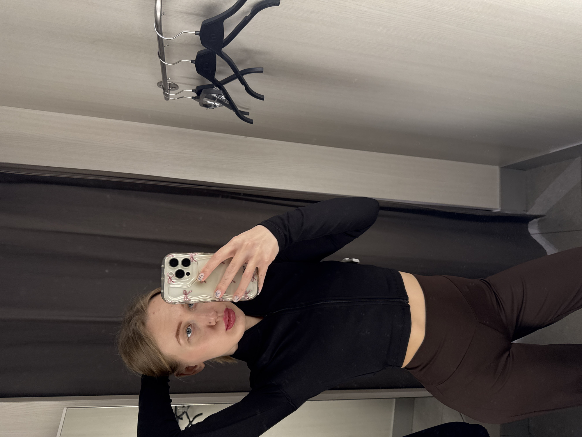 A female subject is depicted in a retail fitting room located in Ovoshchnoy, Russia. The subject, a Caucasian female with blonde hair, blue eyes, a nose piercing, and red lipstick, is wearing a black long-sleeved zipped crop top and dark brown high-waisted pants. She is positioned to take a mirror selfie, holding a light-colored iPhone with a clear case decorated with pink bow motifs in her right hand. Her left arm is raised, with her hand positioned near the back of her head. A partial reflection of the subject is visible in a mirror on the left side of the frame. The fitting room features dark fabric (likely a curtain) to the left, light-colored, textured walls, and a silver-toned metal rod with three black plastic clothing hangers mounted on the right wall.