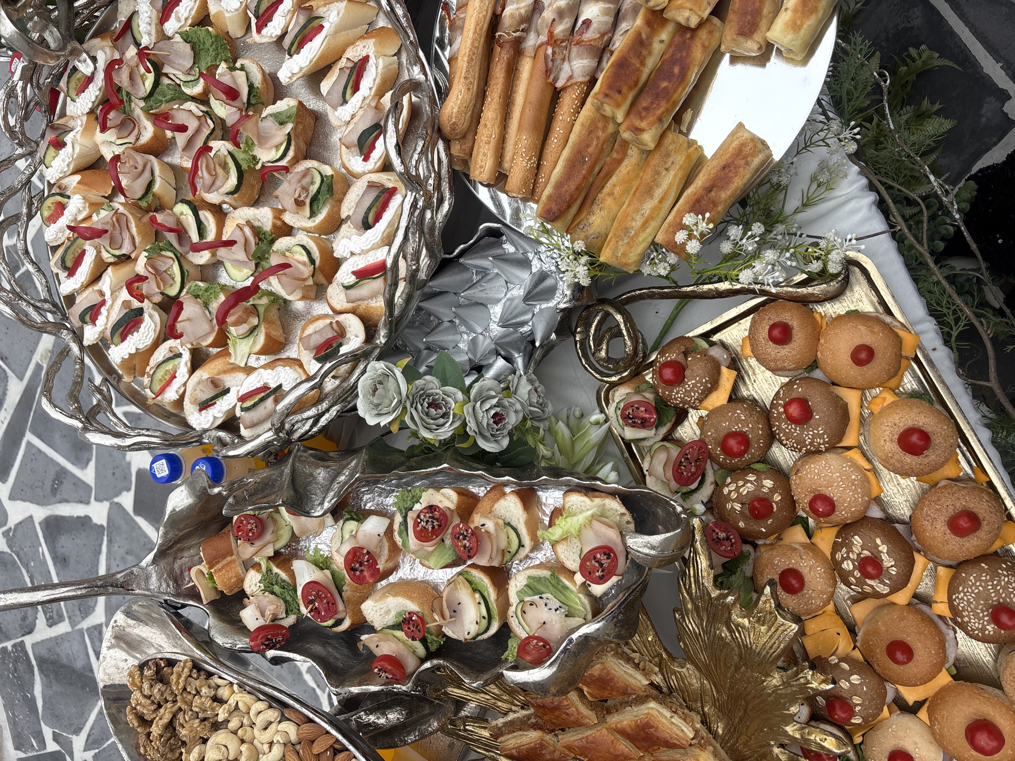 The image shows an assortment of food arranged on ornate silver and gold platters. The platters are placed on a surface with floral decorations.
The food includes small sandwiches with various toppings, such as cream cheese, ham, cucumber, and red peppers. There are also breadsticks, some wrapped in bacon, and small, filled pastries. In another section, there are miniature burgers with a red cherry on top, placed on a gold tray. Another platter has a mix of nuts, including cashews, almonds, and walnuts.
The overall impression is of a catered event or a buffet-style display of appetizers. Without additional context, it is impossible to confirm the specific location in Yerevan, Armenia, or any event that might be occurring.