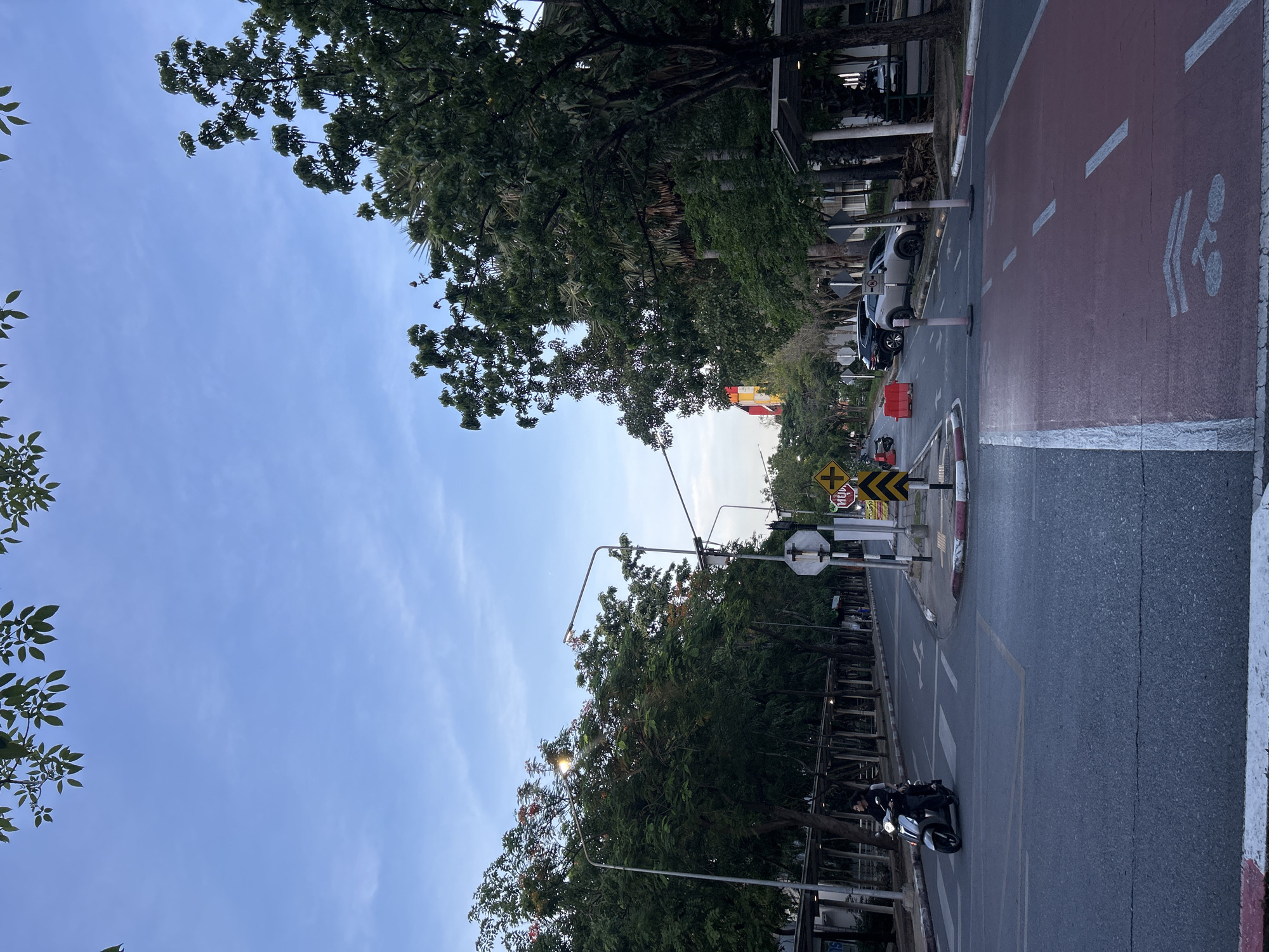 The image depicts a street scene in Tha Khlong Town Municipality, Thailand, during what appears to be late afternoon or early evening. The sky is a pale blue with scattered clouds. Lush green trees line both sides of the road, some with orange-red flowers visible.
In the foreground, a paved road runs through the scene, with a dedicated red-colored bike lane marked on the right side. A person is riding a scooter heading towards the viewer on the main road, passing over a pedestrian crossing marked with white stripes.
Further down the road, there are several parked cars, including a silver SUV. Traffic signs are visible, including a yellow diamond-shaped sign with a black arrow and a stop sign with Thai text. Orange traffic cones and barriers are also present. On the left side of the road, a covered walkway or arcade runs alongside buildings, with more trees and vegetation. Streetlights are mounted on poles along the road. The overall environment suggests an urban or suburban area with ample greenery.