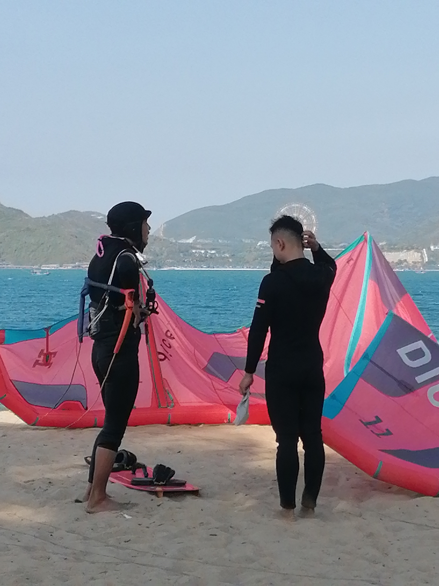 Two people are standing on a sandy beach in Nha Trang Ward, Vietnam. The person on the left is wearing a black wetsuit and helmet, and a harness over the wetsuit. They are barefoot and standing near a pink and purple kiteboard on the sand. The person on the right is wearing a black wetsuit and is barefoot. They are holding a white cloth in their left hand and touching the back of their head with their right hand. There are two large, deflated pink and purple kites lying on the sand behind them. In the background, there is the ocean, and in the distance, there are green mountains and buildings. There is a ferris wheel to the right of the mountains. The sky is a light blue, indicating it is daytime.