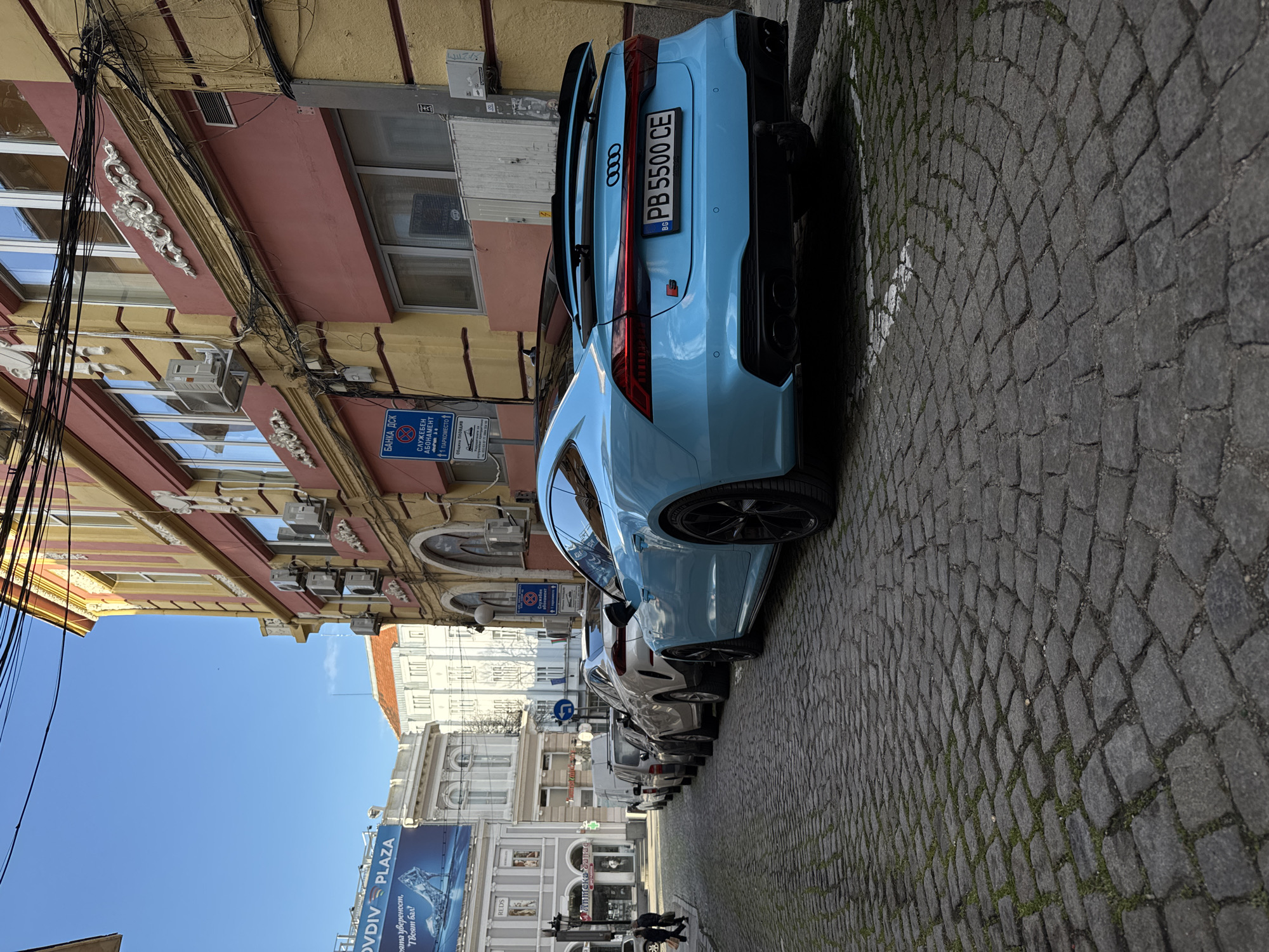The image shows a row of parked cars on a cobblestone street in Plovdiv, Bulgaria. The car closest to the camera is an ice-blue Audi RS with a license plate reading "PB 5500 CE." A sign for "BANKA ДСК" and "СЛУЖБЕН АБОНАМЕНТ" (Service Subscription) with parking information is affixed to the building next to the Audi. Above the cars is a view of the sky and the facades of the surrounding buildings. The "Plovdiv Plaza" advertisement is visible in the background. A couple of people are walking on the street towards the background.