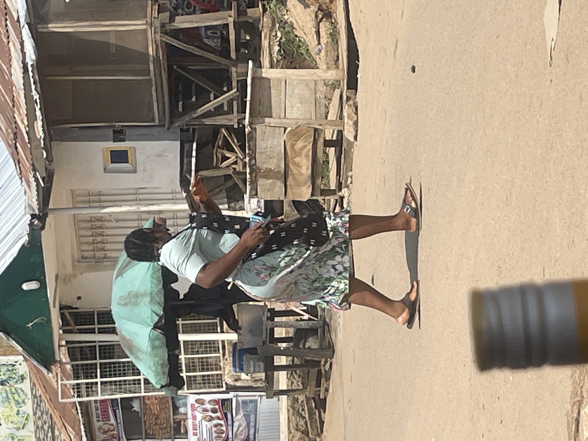 The image depicts a street scene in Abuja, Nigeria, likely during the daytime. The main subject is a person standing on a dirt or paved road, holding a phone and possibly taking a picture or video. They are wearing a light blue t-shirt and a floral-patterned skirt, and are barefoot or wearing sandals. A large, light green umbrella is propped open next to them.
In the background, there is a rustic structure made of wood and corrugated metal, suggesting a market stall or a makeshift shelter. This structure appears to have shelves and various items on display, though they are not clearly discernible. Further in the background, there are more buildings and possibly trees or foliage. The ground is a dusty, light brown color.
To the right of the frame, the blurry end of a yellow cylindrical object is visible, which could be a bottle or a tube of some kind.
The overall scene appears to be in a somewhat informal setting, characteristic of a marketplace or a roadside vendor area. The lighting suggests it is daytime, possibly sunny, but no specific weather conditions can be determined. No overt emotions are displayed by the subject. There is some text visible on a sign in the lower part of the image, but it is too blurry to be read.