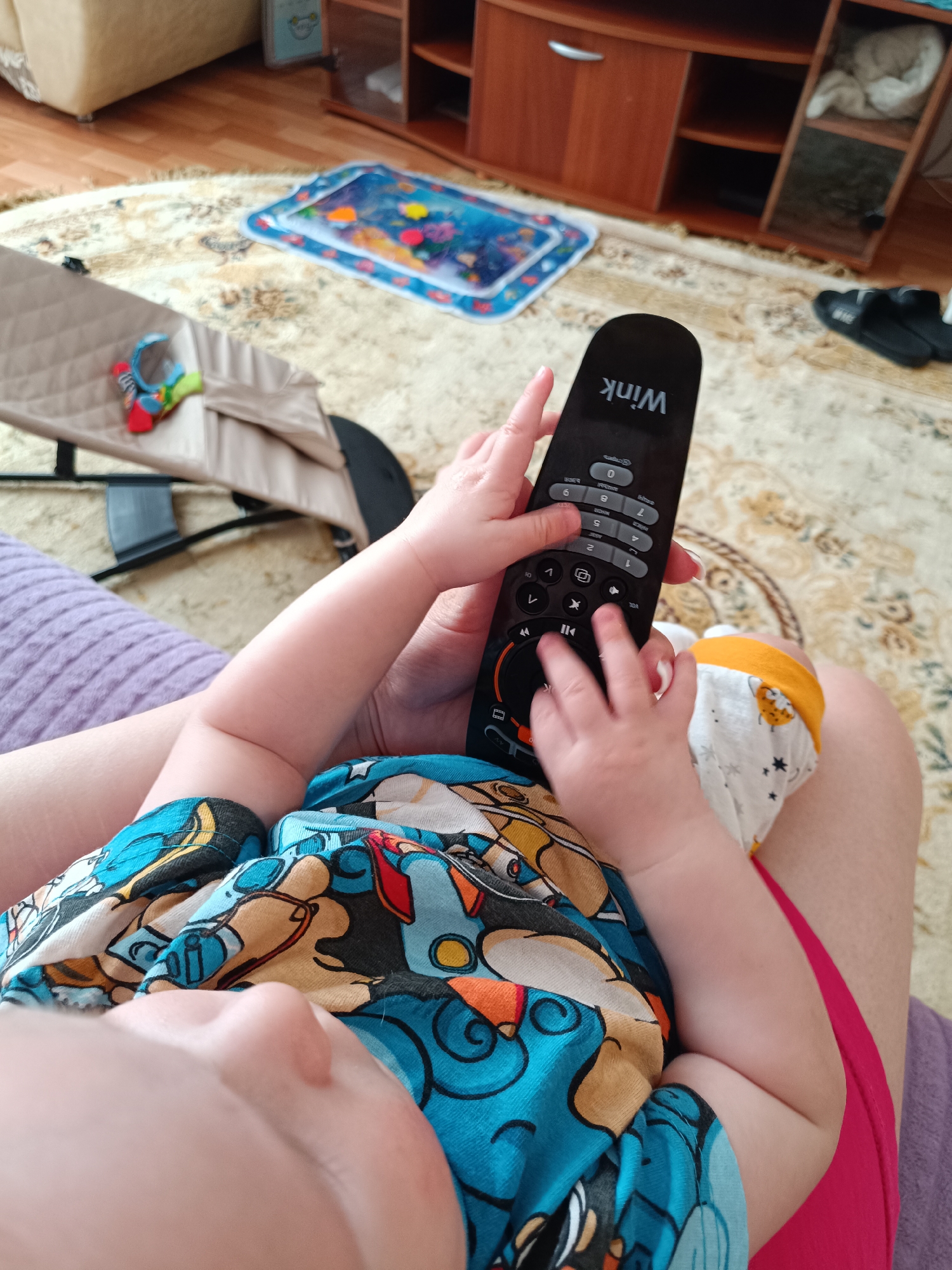 A close-up, eye-level shot shows a baby in a blue and brown cartoon-printed shirt, sitting on the lap of an adult wearing a red skirt. The baby's hands are on a black "Wink" remote control, with the adult's hand visible underneath. The remote has numerous buttons. Background details include a beige patterned rug, a baby bouncer, and a wooden shelving unit. A water-filled play mat sits on the rug. The location of the image is difficult to pinpoint based on the picture. The picture does not display the city of Tobolsk, Russia. The setting appears to be inside a home.