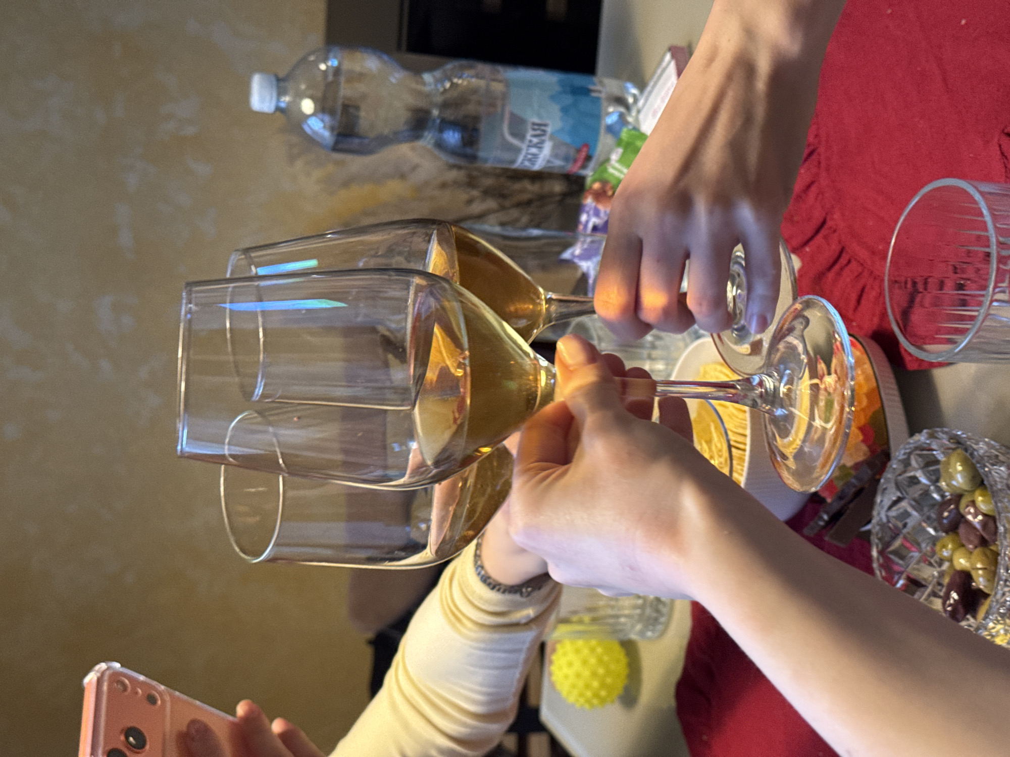 A slightly rotated, close-up shot captures a celebratory event, possibly a toast, taking place indoors in an unspecified city. Two champagne flutes, filled with a golden liquid likely to be champagne or white wine, are being clinked together by two individuals whose arms and hands are visible. One person is wearing a light-colored, long-sleeved garment, while the other has bare arms. A smartphone with a peach-colored case is visible in the lower left, presumably used to capture the moment.
The background includes a table covered with a red cloth. Visible on the table are a bottle of water with Russian labeling, a glass tumbler, a decorative bowl filled with assorted candies, and a small yellow spiky ball, possibly a toy or decoration. The surrounding environment is blurred, revealing a light-colored wall with a textured surface in the background and a dark rectangular shape above the bottle.