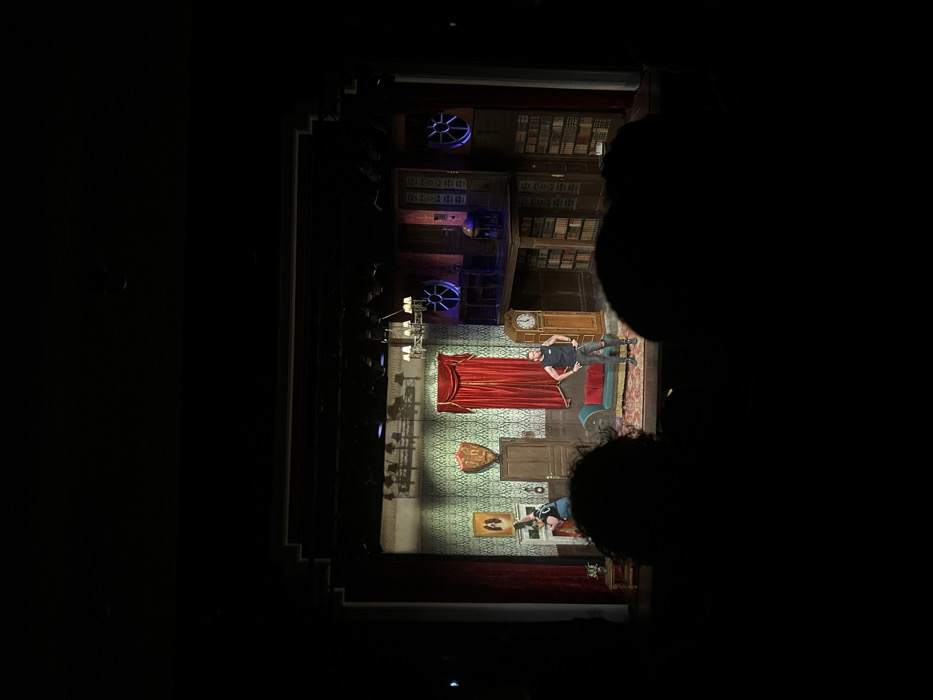 A theater stage set displays a whimsical scene. A man in a black shirt and dark pants lies on a rug next to a red chaise lounge. Above, a large grandfather clock stands against a bookshelf-lined wall. Below, a second actor leans against a lower section of the set. A chandelier hangs above, illuminating the patterned wallpaper. Stage lights with fan-like coverings are mounted higher in the background. The view is partially obstructed by blurry heads in the foreground.